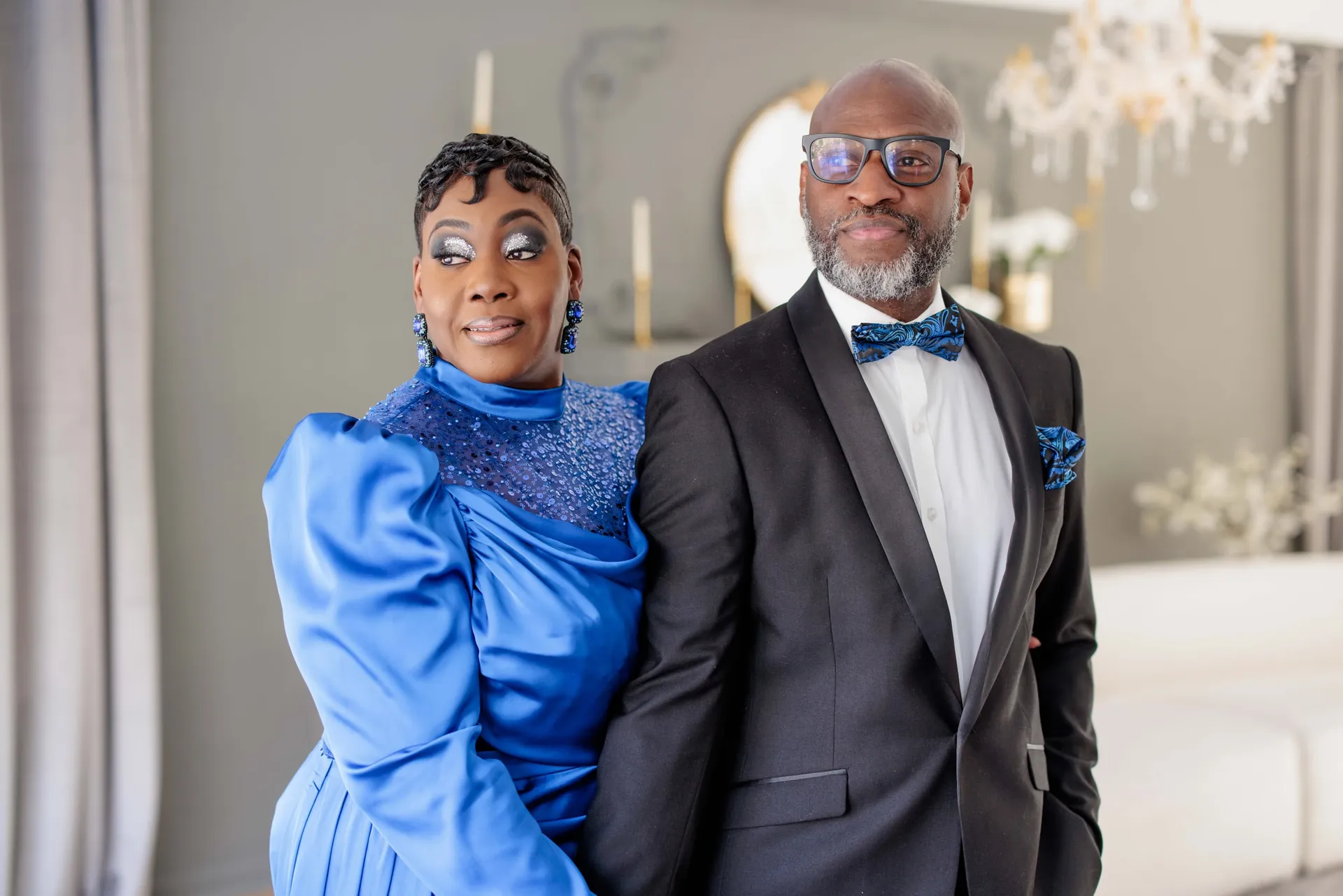 An anniversary couple in royal blue and a black tuxedo posing in a bright white room.