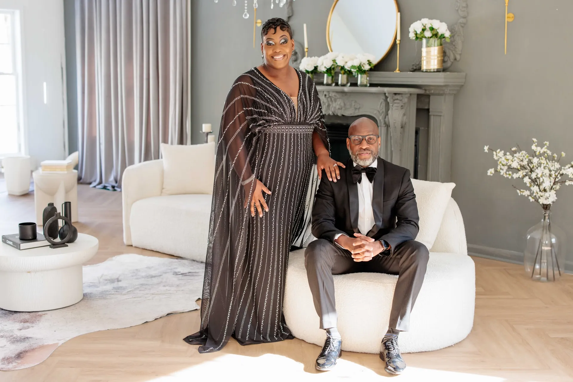 An anniversary couple in a bright styled lounge, she standing beside his armchair in evening wear.