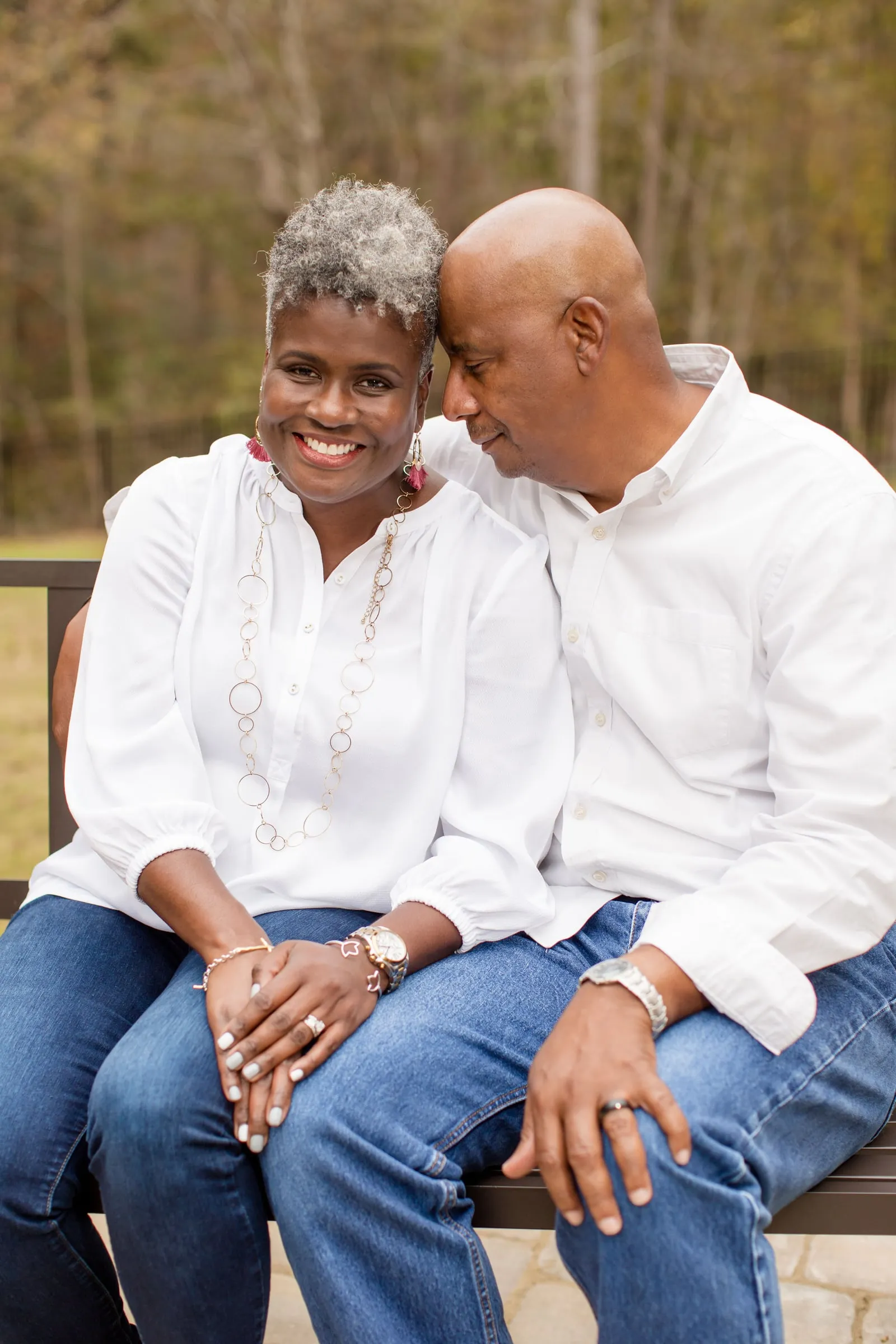 A couple in white shirts and jeans leaning their heads together on a park bench.