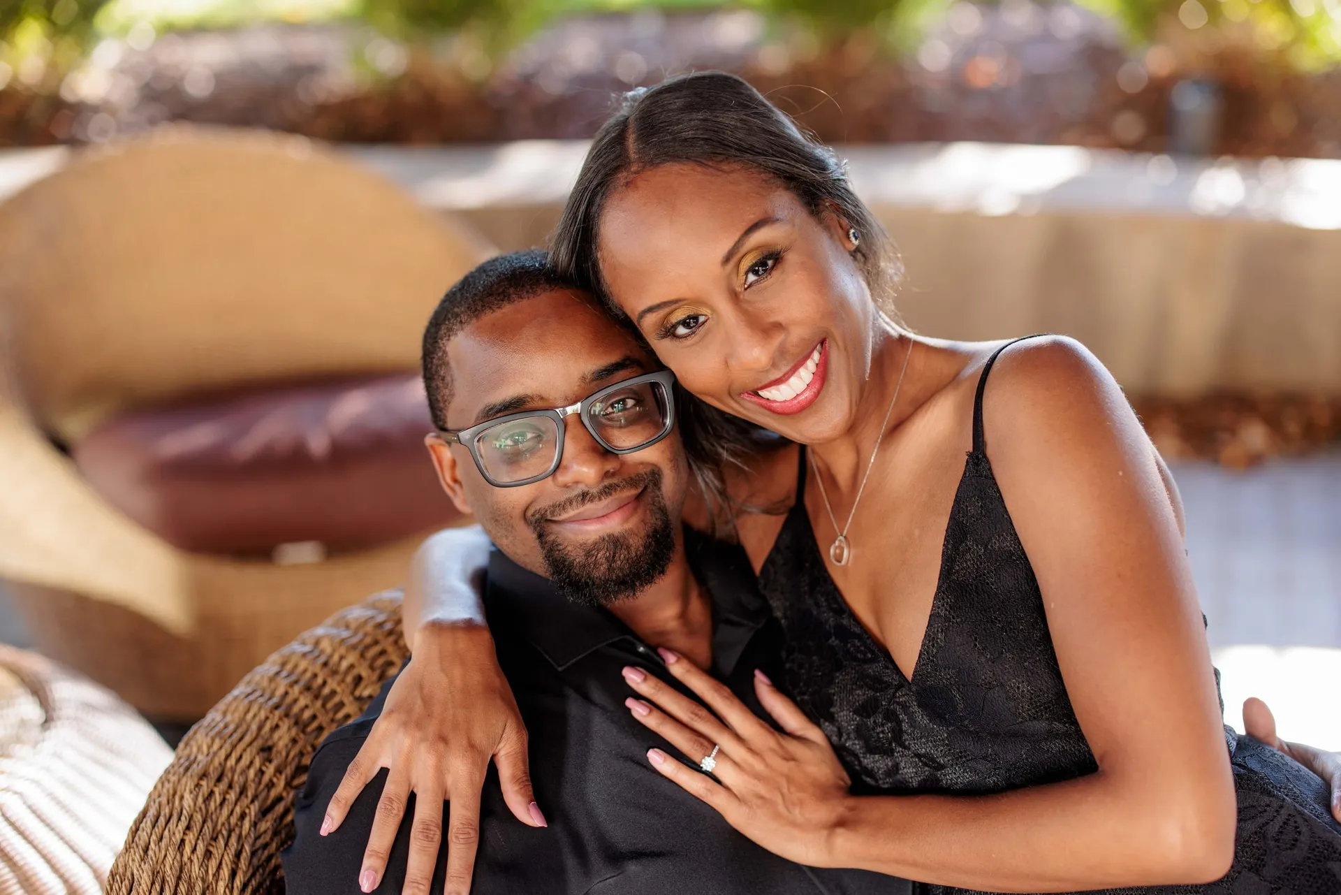 An engaged couple cheek to cheek in a wicker chair, her ring hand on his shoulder.