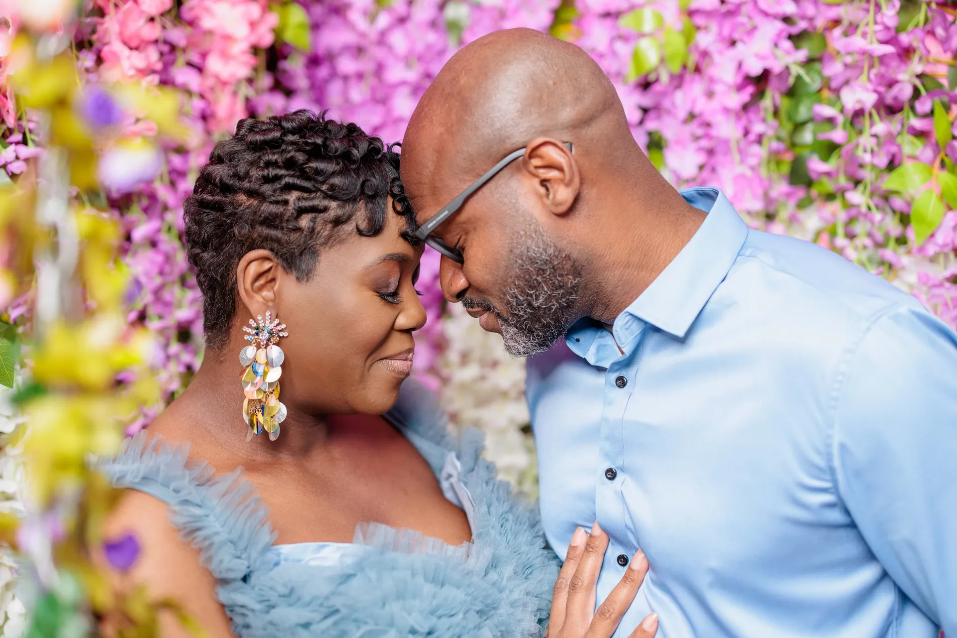 An expectant couple forehead to forehead beneath pink blossoms, she in a blue tulle dress.