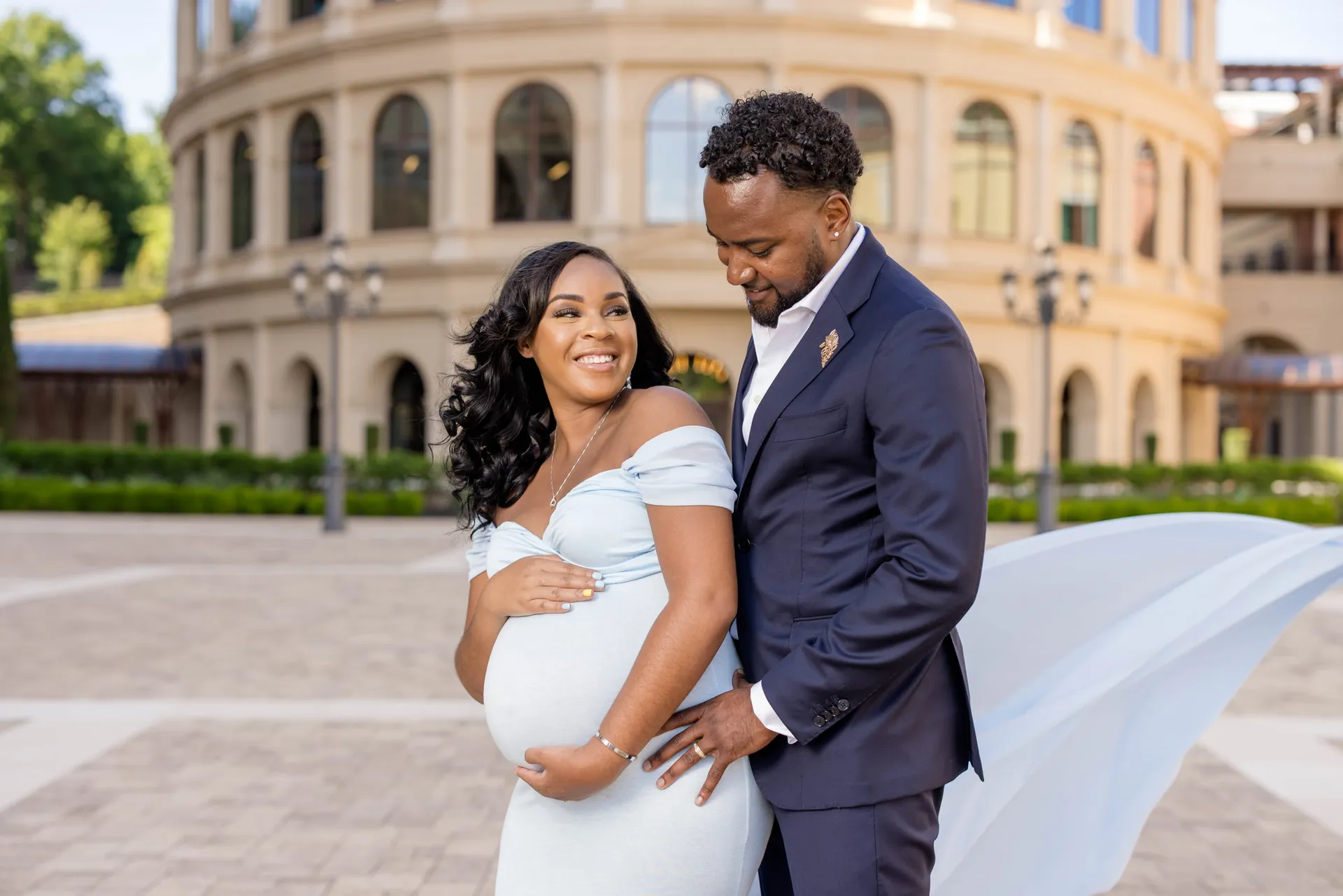 An expectant mother in pale blue smiling as her partner in navy looks at her bump.