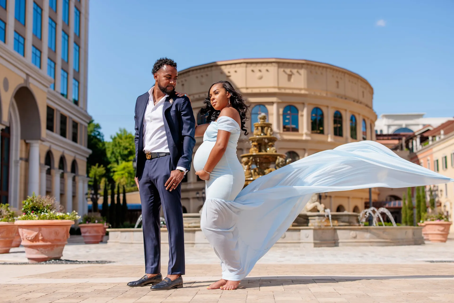 An expectant couple on a sunlit plaza, her pale blue gown sweeping across the stone.