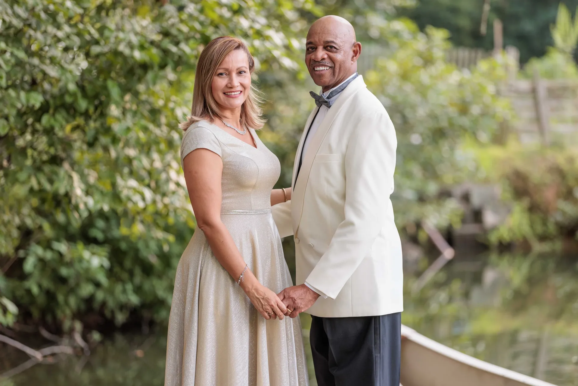 A couple holding hands at the water's edge in a white dinner jacket and champagne dress.