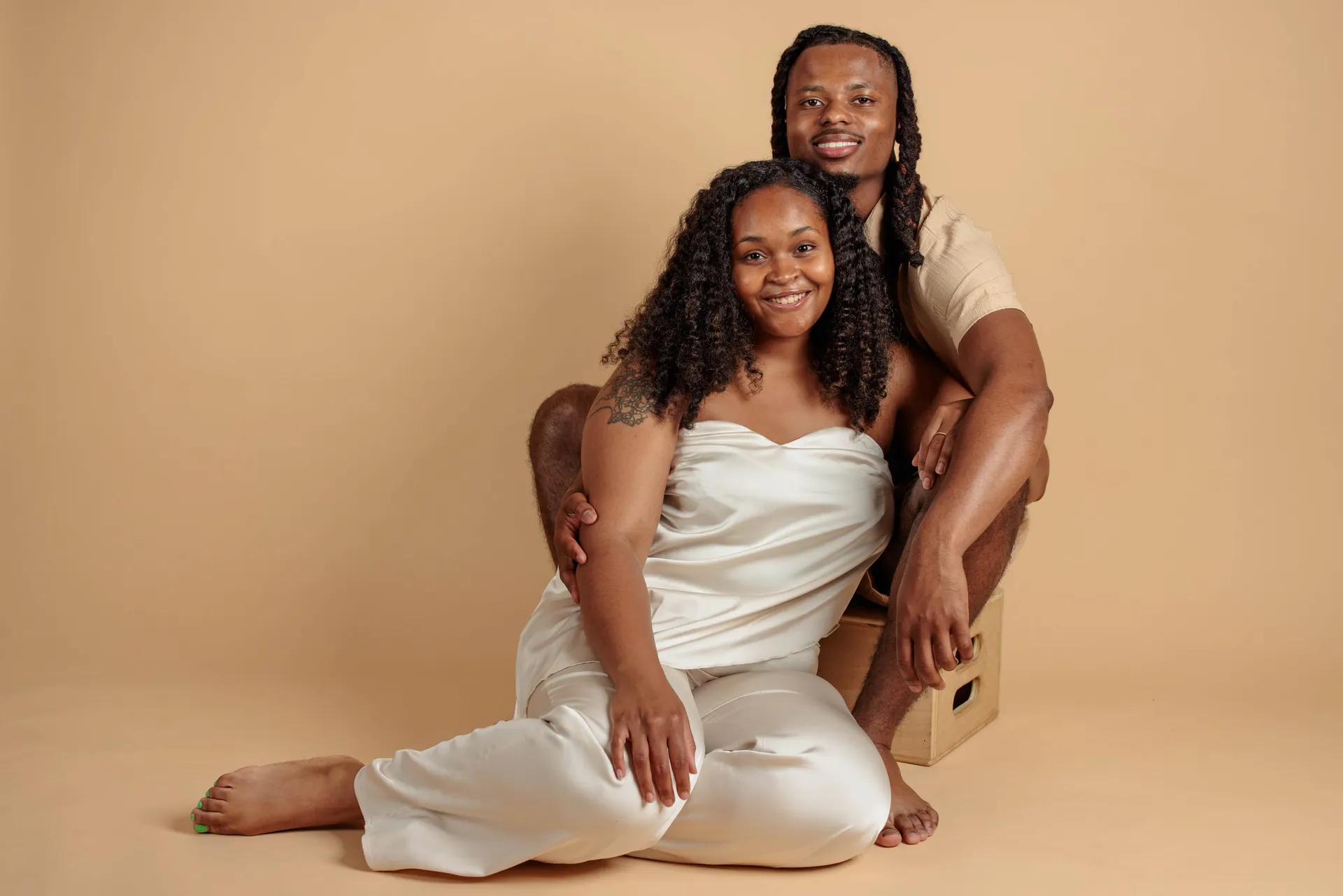 A couple in white seated together on the floor of a warm beige studio backdrop.