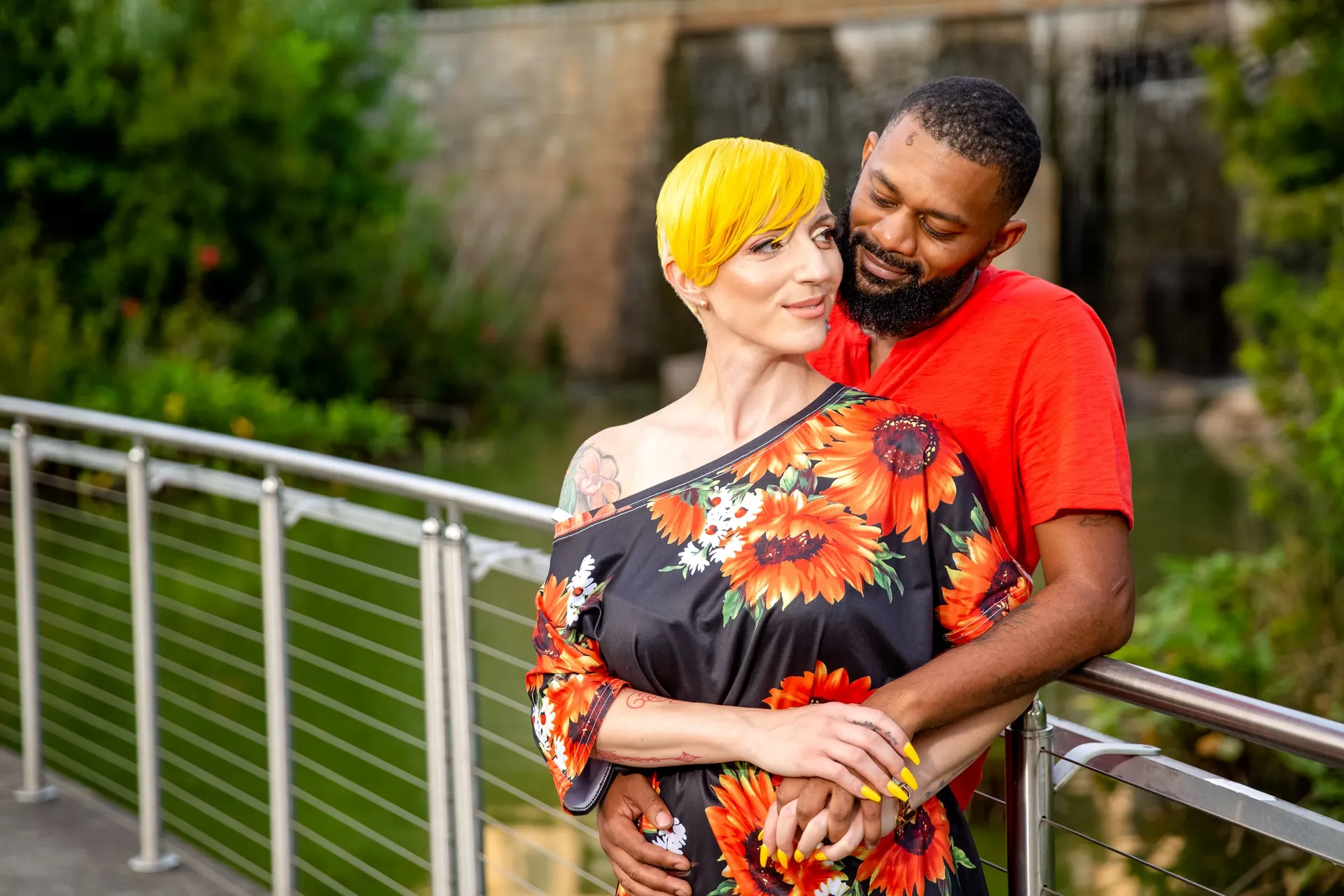A couple in a sunflower dress and red tee leaning at a railing beside a waterfall.