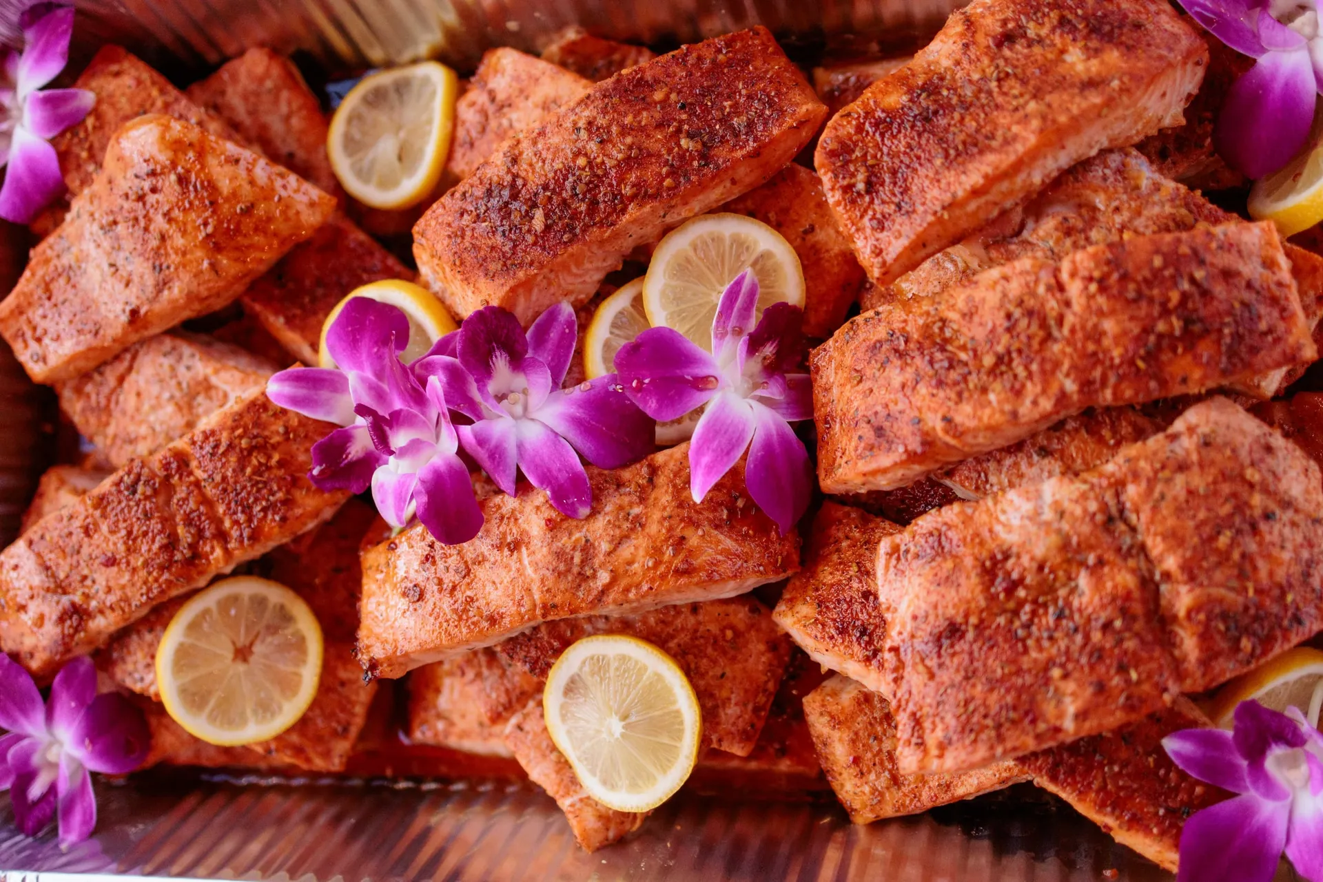 Seasoned salmon bites garnished with lemon slices and purple orchids on a serving board.