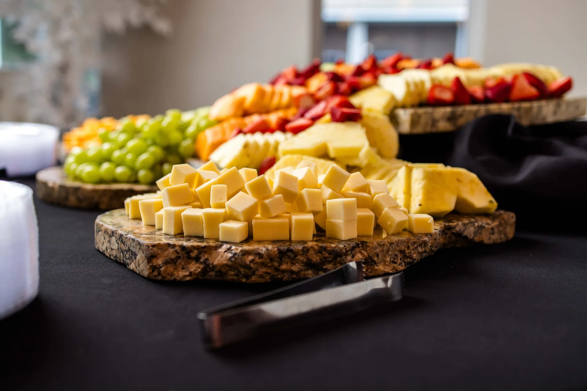 A stone board of cheese cubes, grapes, pineapple and berries on a black-draped table.