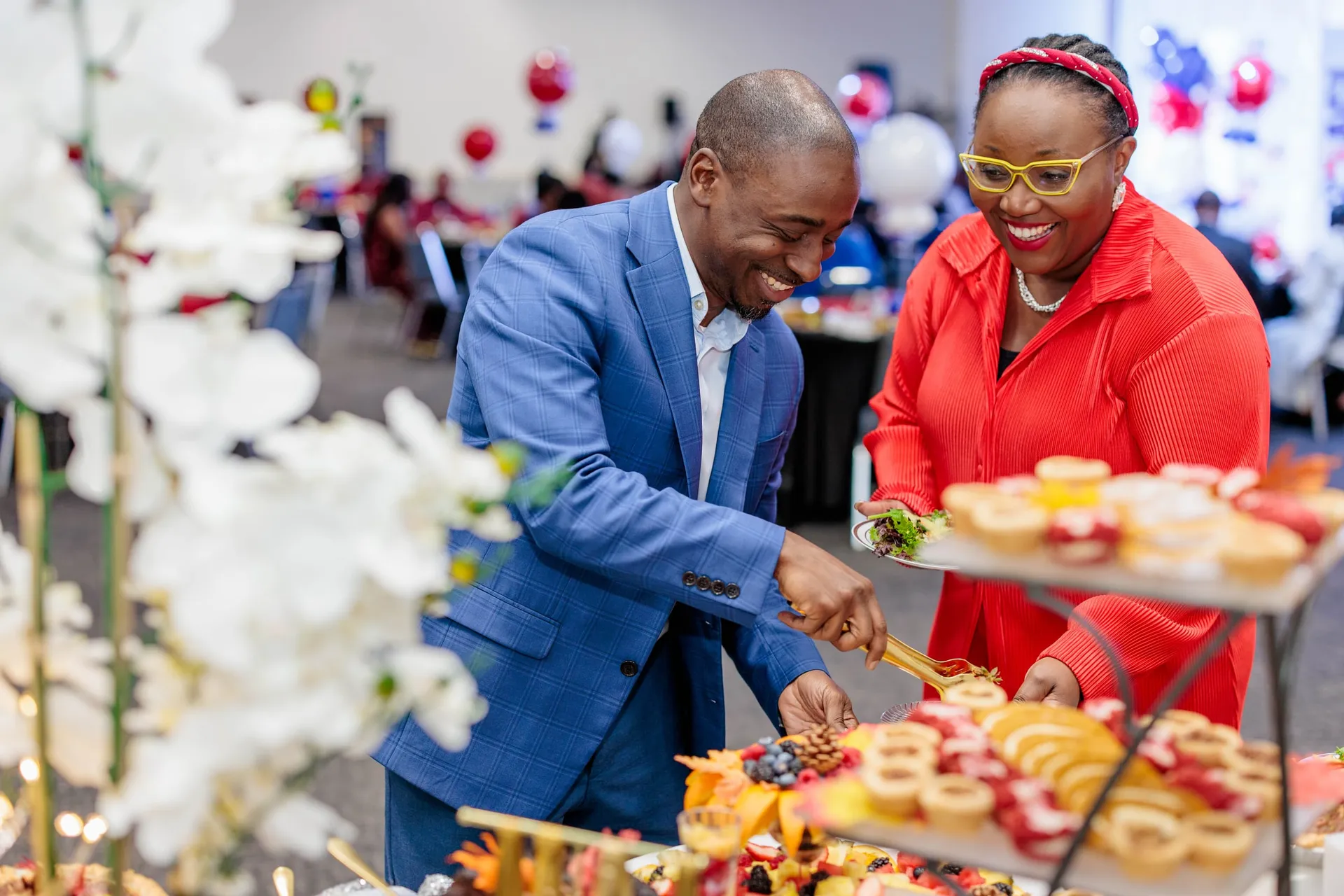 Two guests laughing as they fill their plates at a banquet buffet beside white orchids.