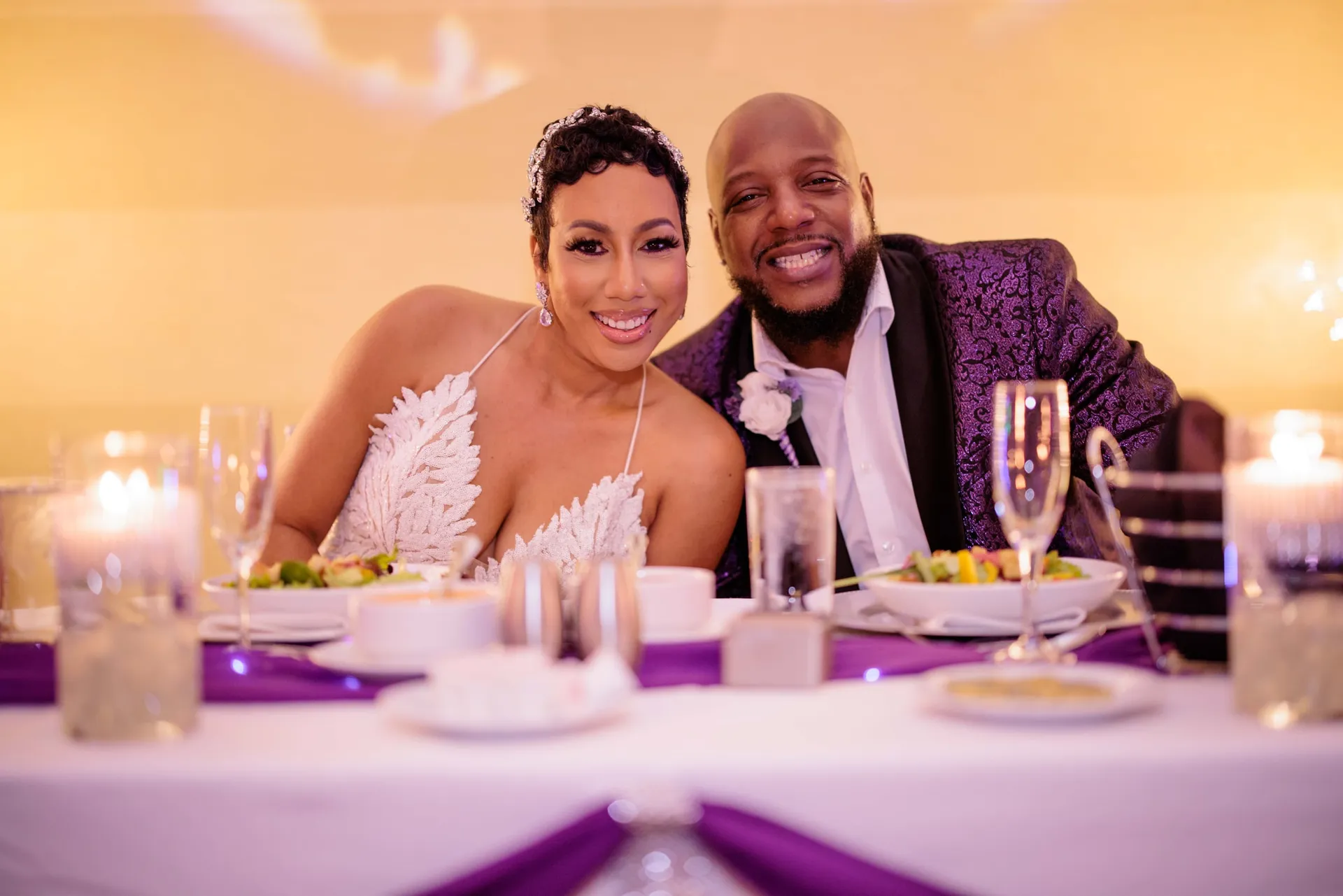 A couple in white lace and a purple brocade jacket smiling at their candlelit head table.