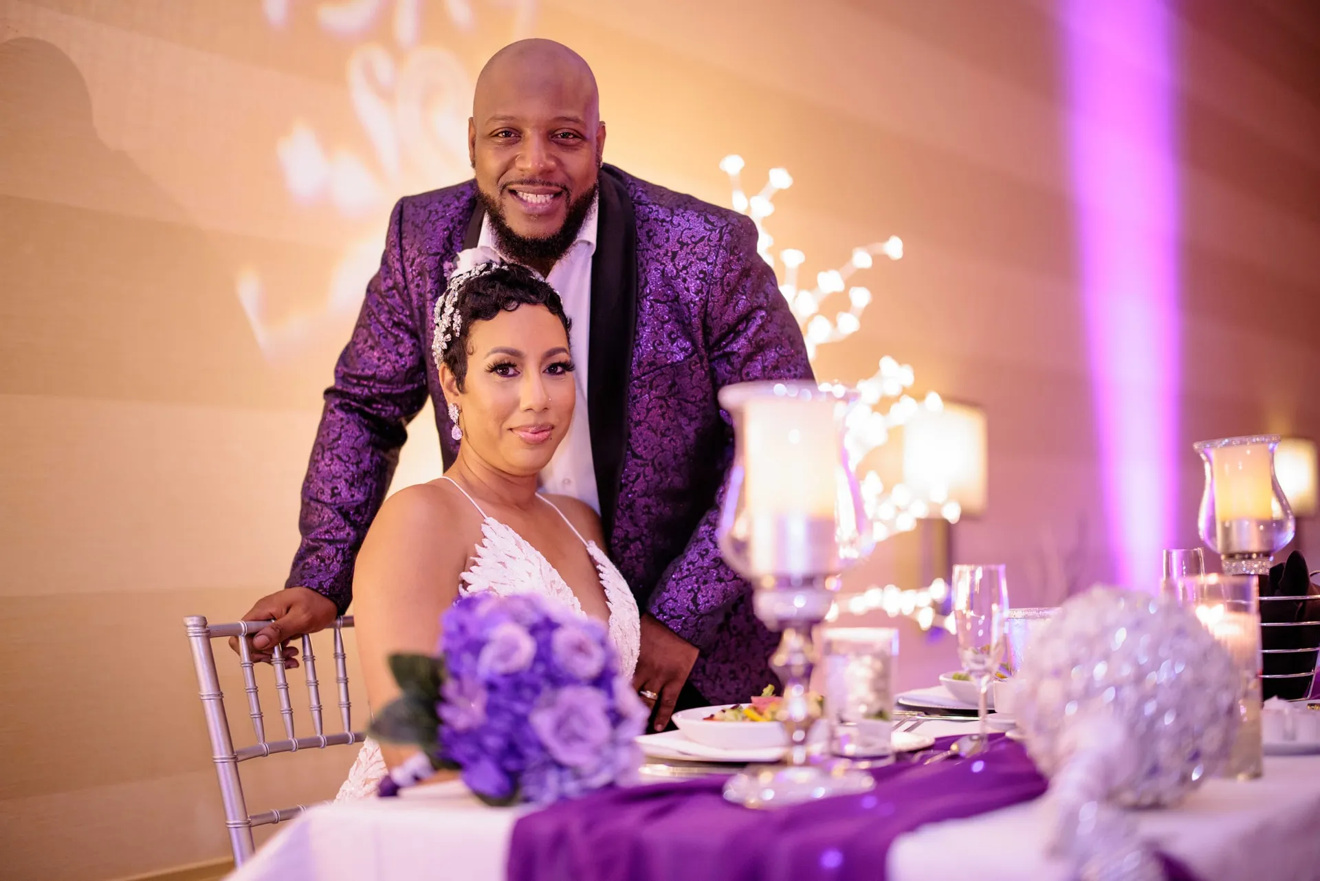 A couple at a purple-draped reception table, he standing behind her chair in a brocade jacket.
