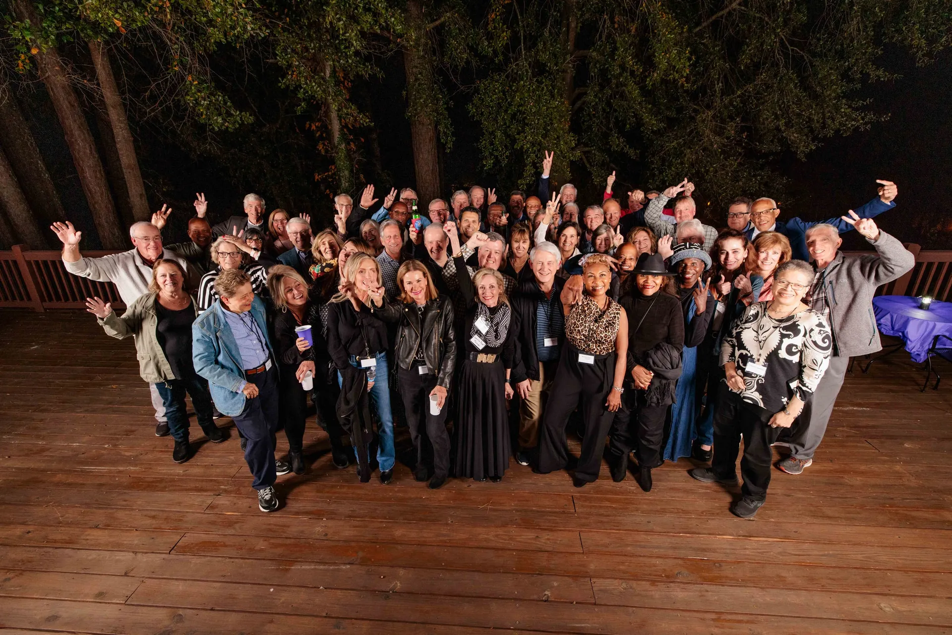 A class reunion crowd waving up at the camera from a wooden deck at night.