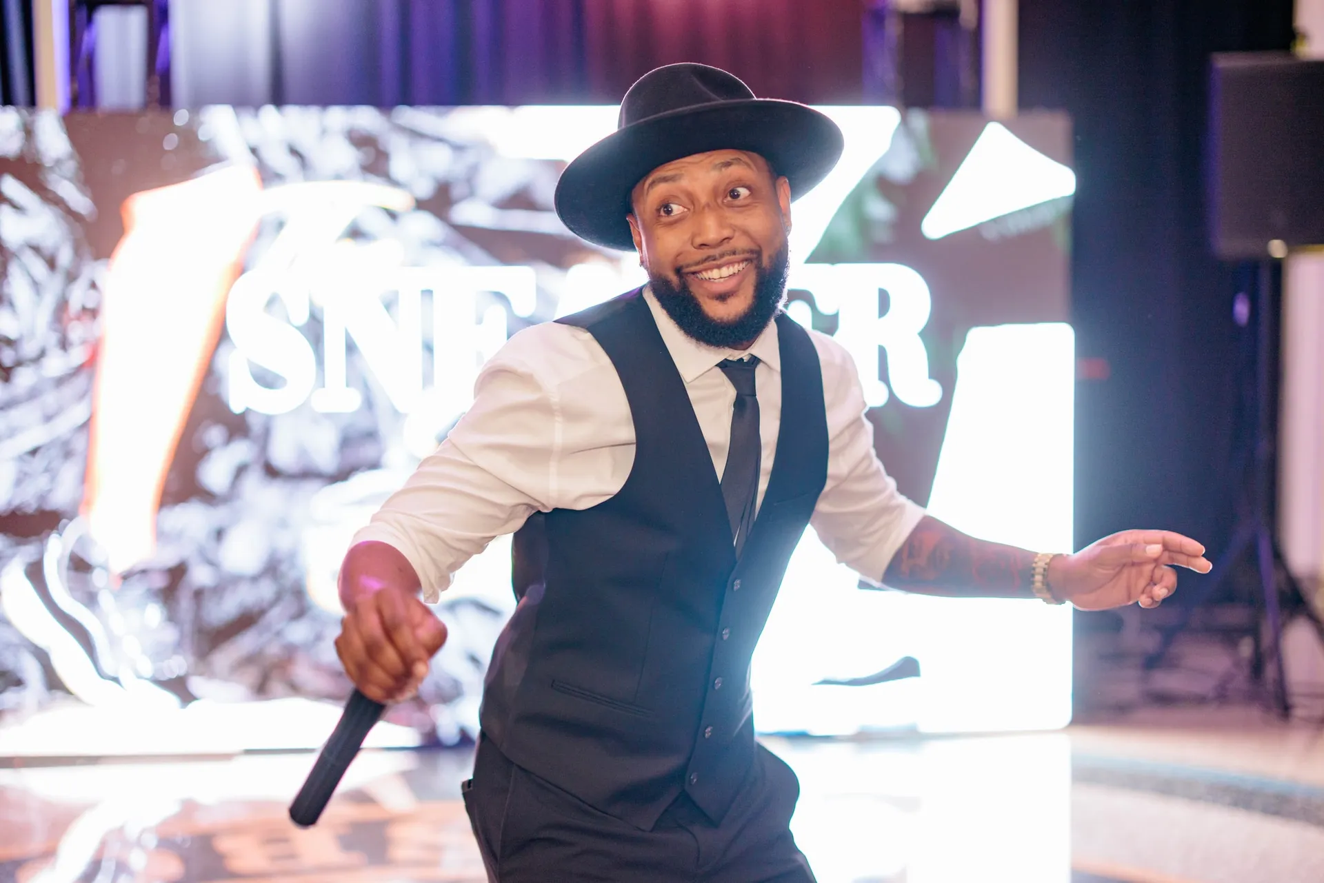 The host in a hat and waistcoat dancing with a microphone before the sneaker ball sign.