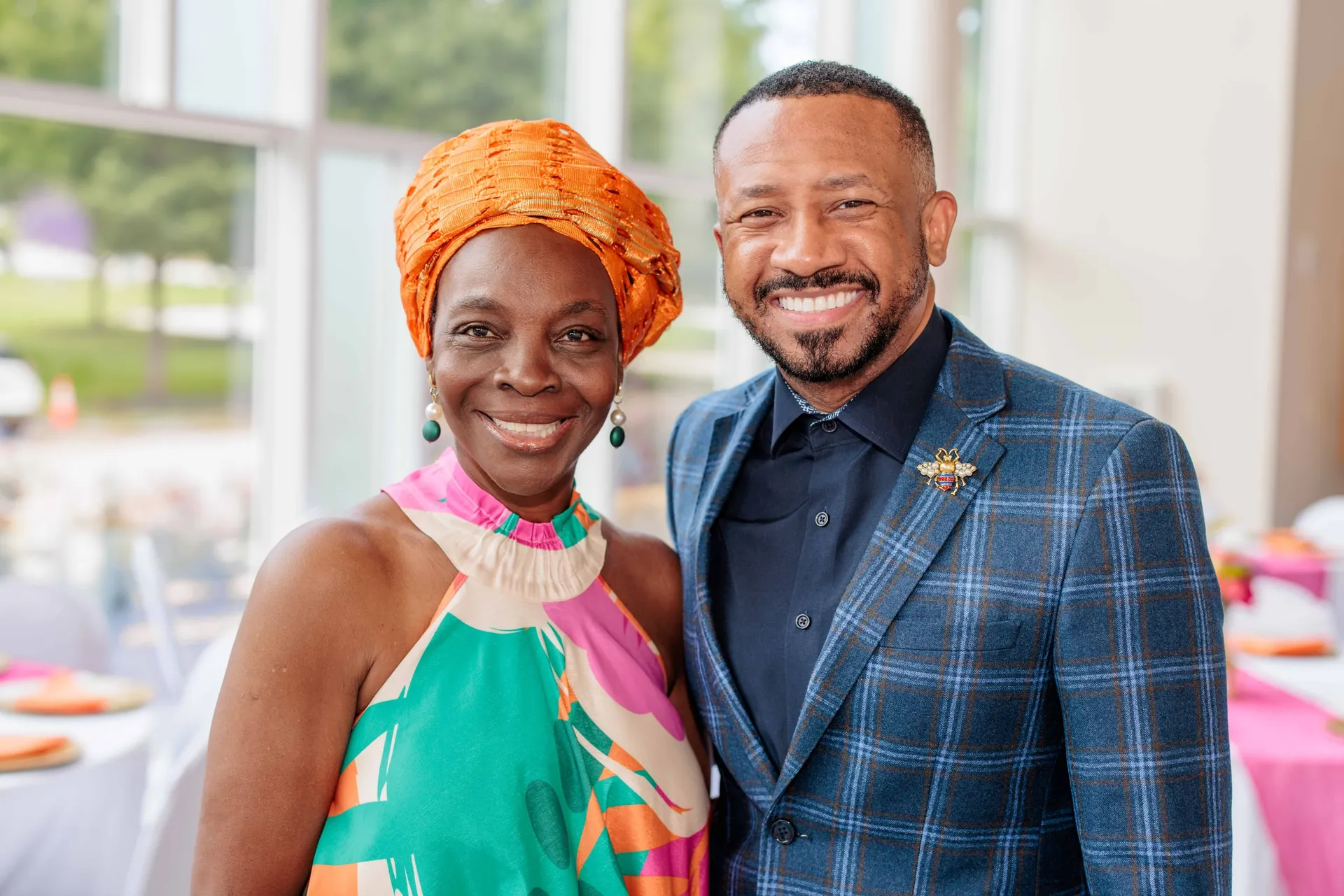Two smiling guests, one in an orange headwrap and tropical dress, one in a plaid blazer.