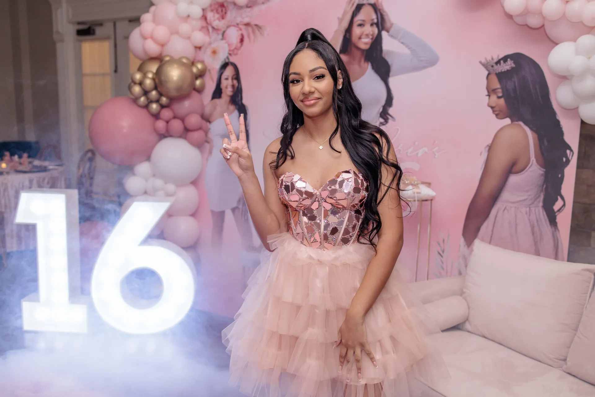 The birthday girl in pink tulle flashing a peace sign beside a glowing 16 and balloons.