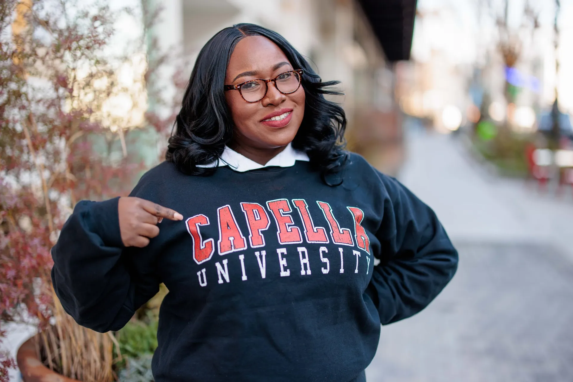 A graduate pointing proudly at her Capella University sweatshirt on a city sidewalk.
