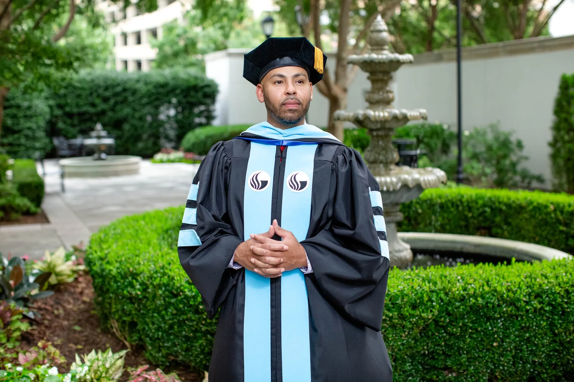 A doctoral graduate in black and light blue regalia in a formal garden beside a fountain.
