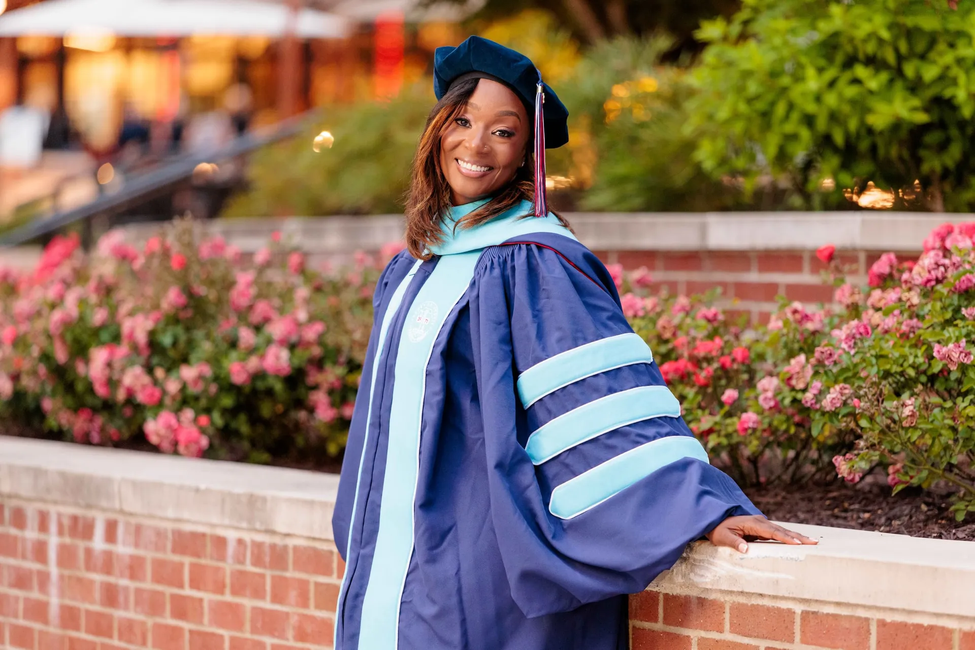 A doctoral graduate in navy regalia leaning on a brick wall beside pink roses at dusk.