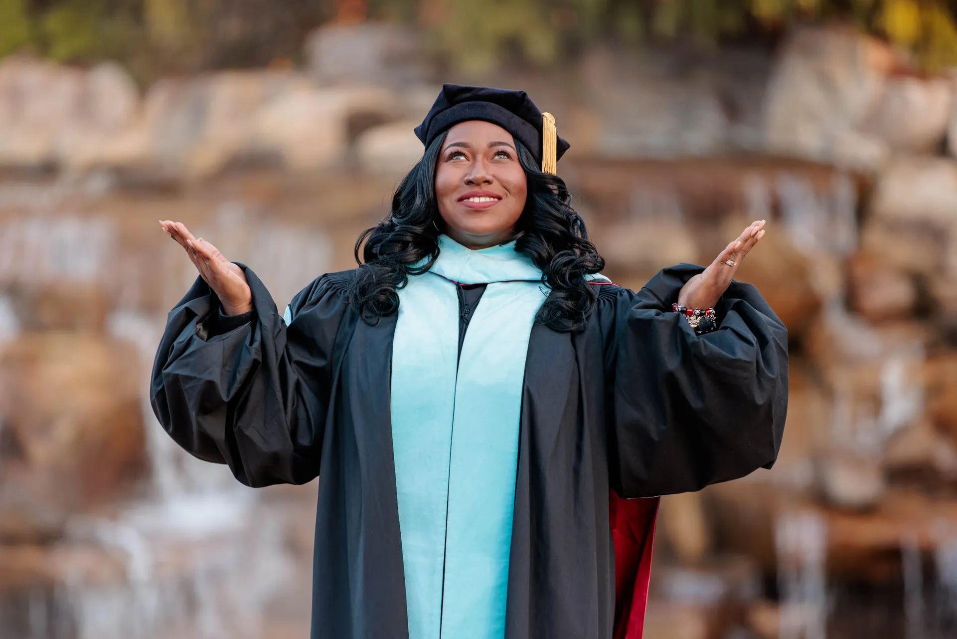 A doctoral graduate in black and light blue regalia opening her arms before a rocky waterfall.