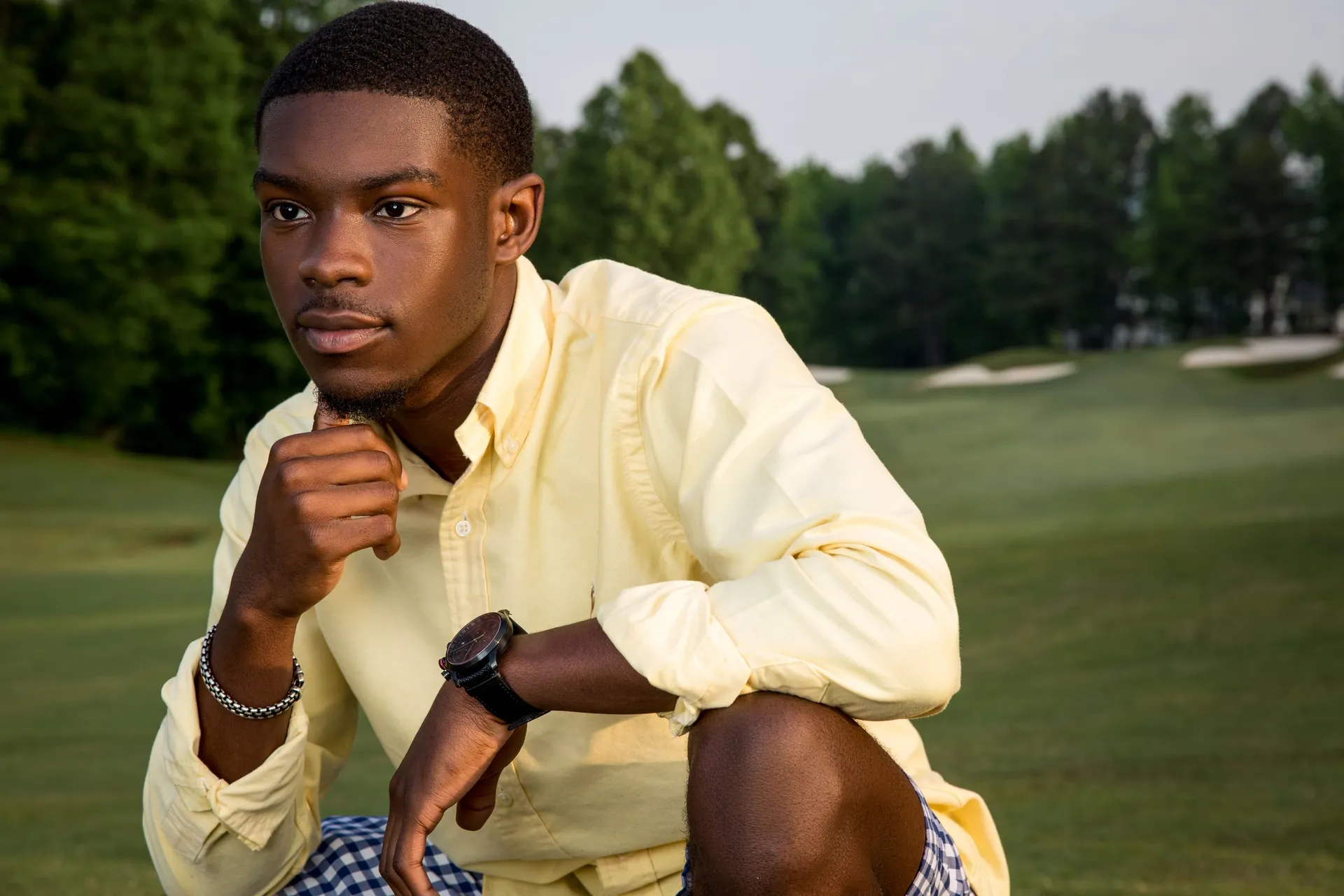 A senior in a yellow shirt crouched on a golf fairway, fist under his chin.