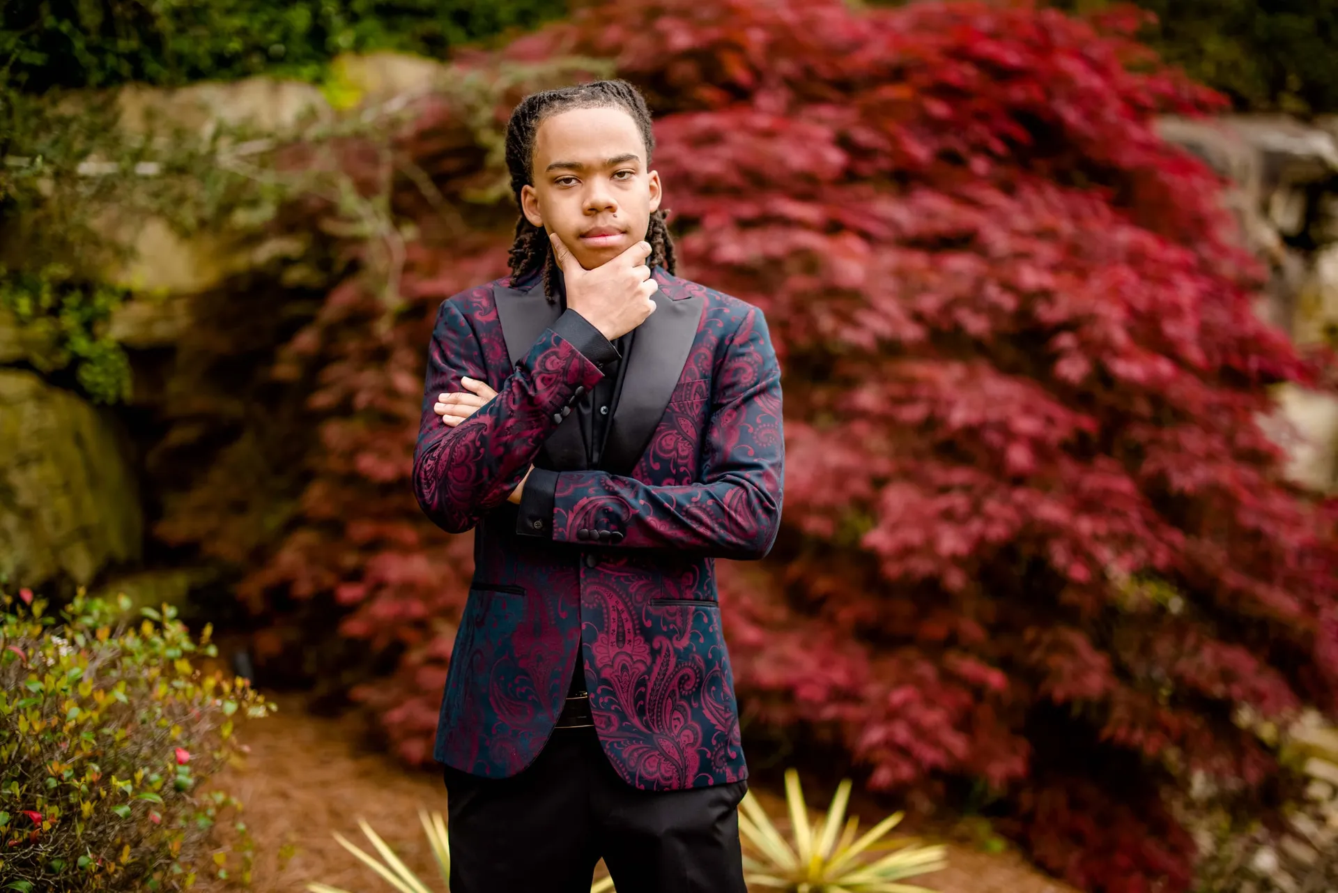 A senior in a burgundy paisley blazer standing thoughtfully beside a red Japanese maple.