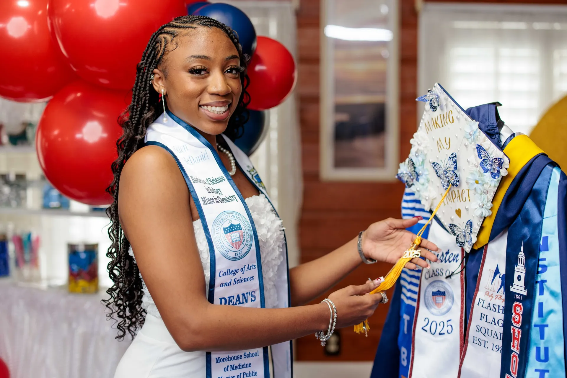 A graduate in white holding her decorated cap beside a rack of honor stoles and balloons.