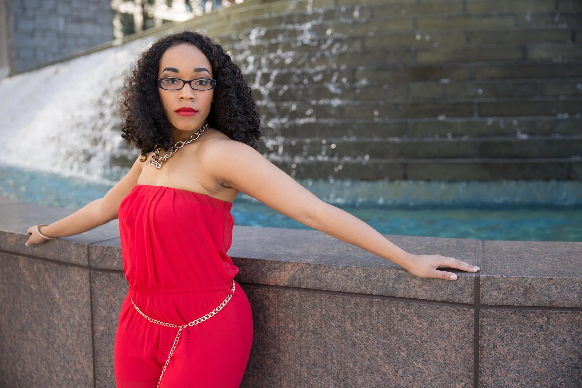 A senior in a red jumpsuit leaning along a granite wall before a stepped fountain.