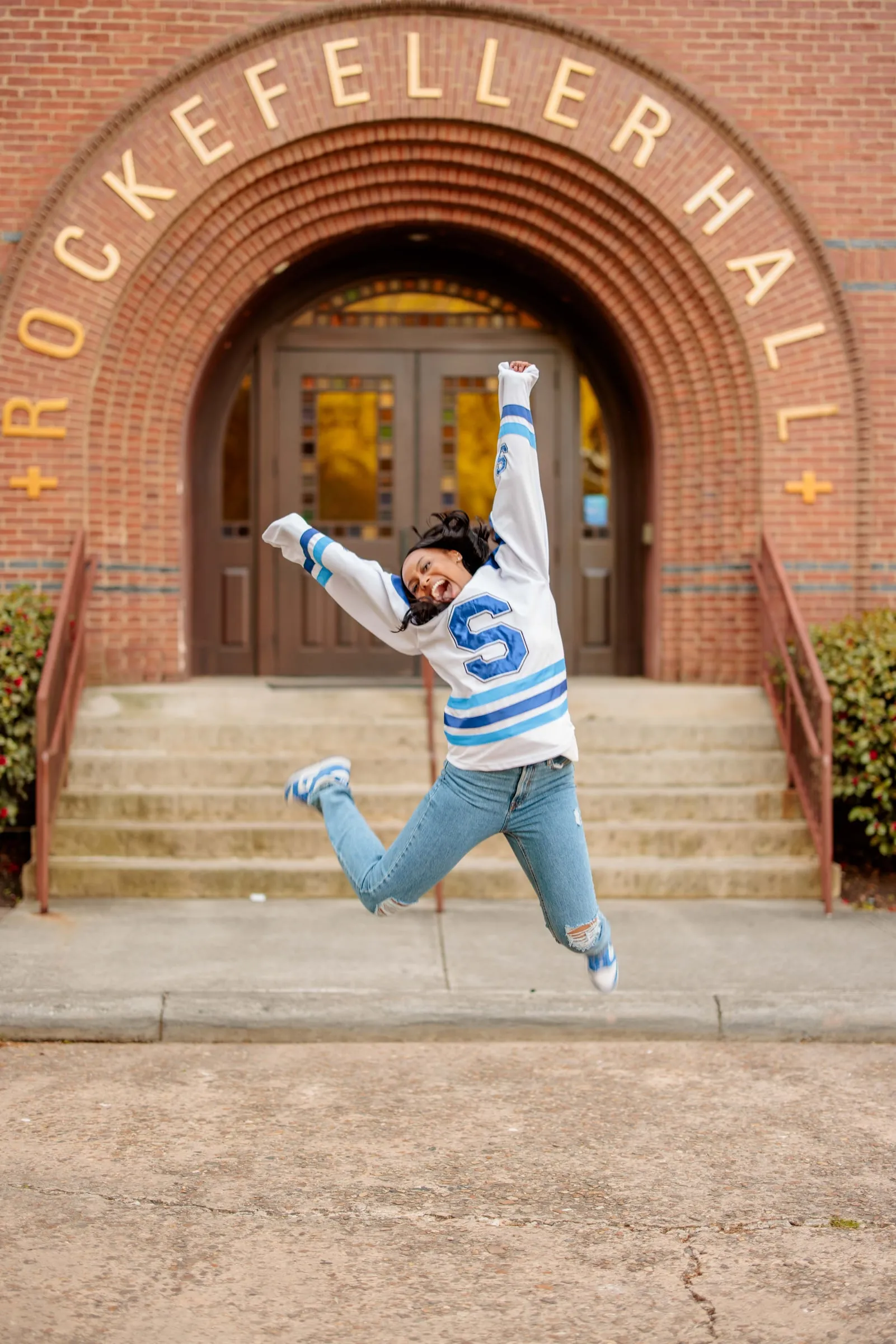 A graduate in a Spelman sweater leaping for joy before the brick arch of Rockefeller Hall.