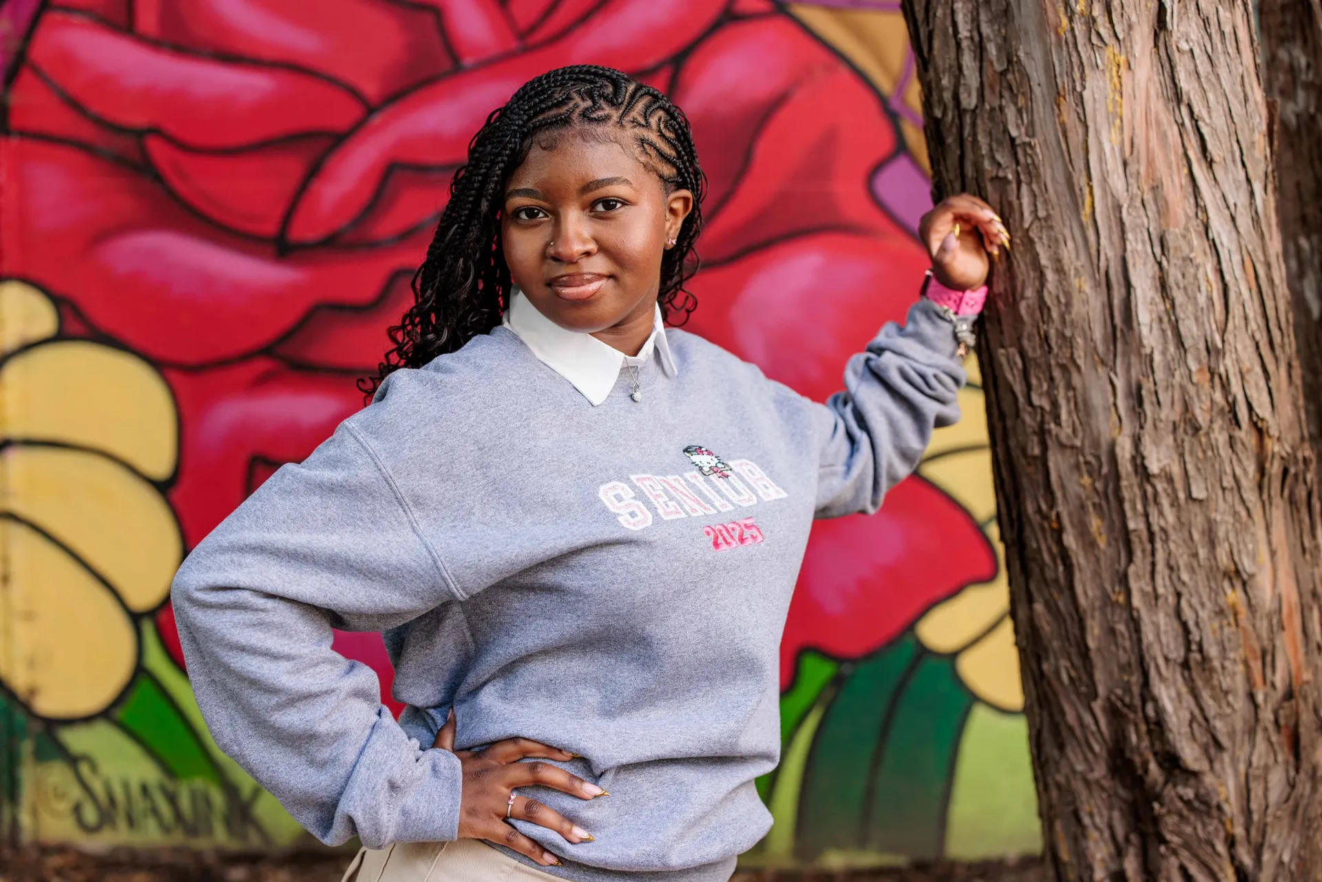 A senior in a grey Senior 2025 sweatshirt beside a tree before a giant rose mural.