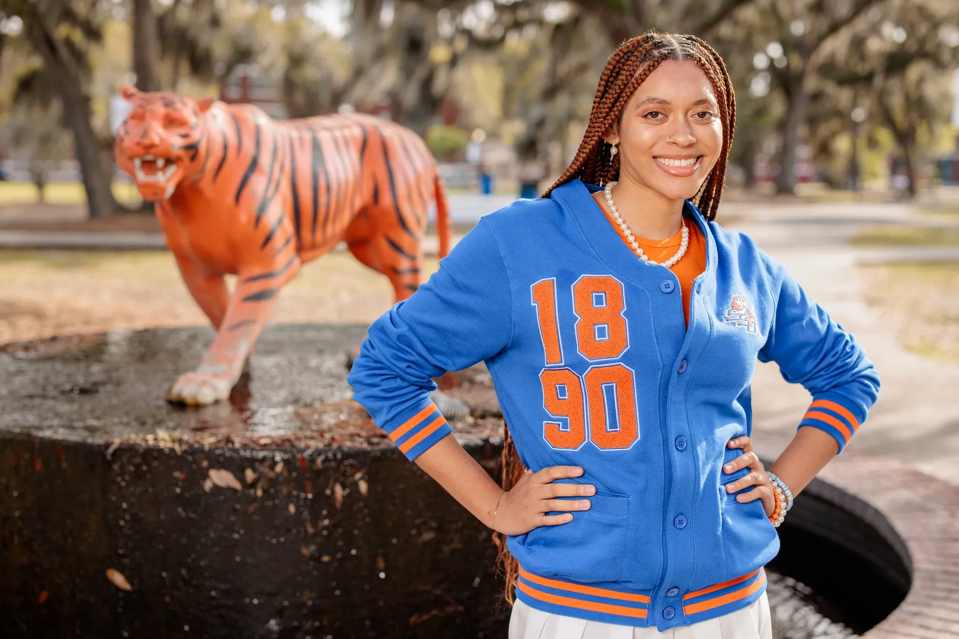 A graduate in a blue and orange letterman jacket before a tiger statue under Spanish moss.