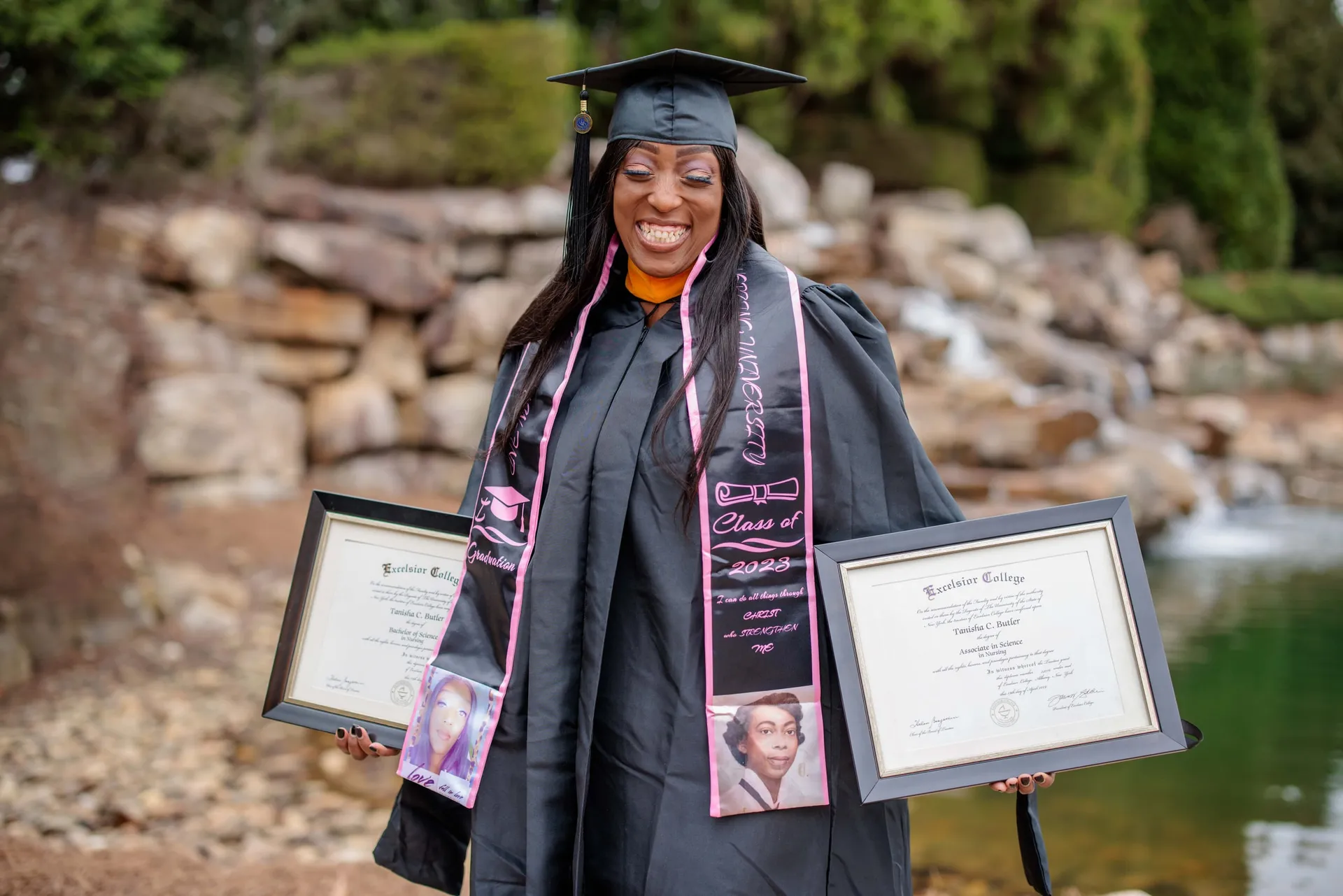 A graduate in cap and gown holding up two framed diplomas before a rocky waterfall.