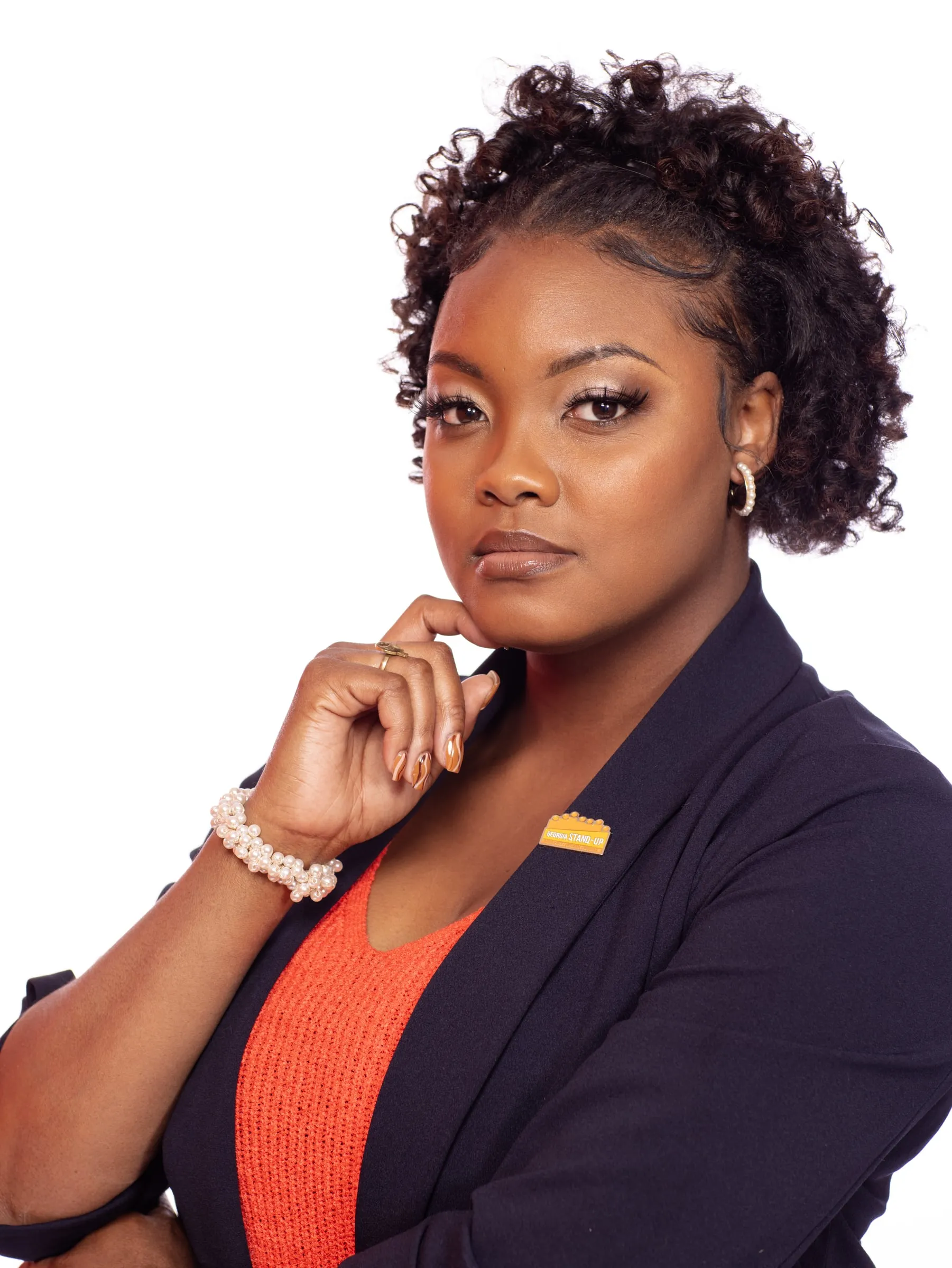A studio headshot in a navy blazer and orange top, hand at her chin.