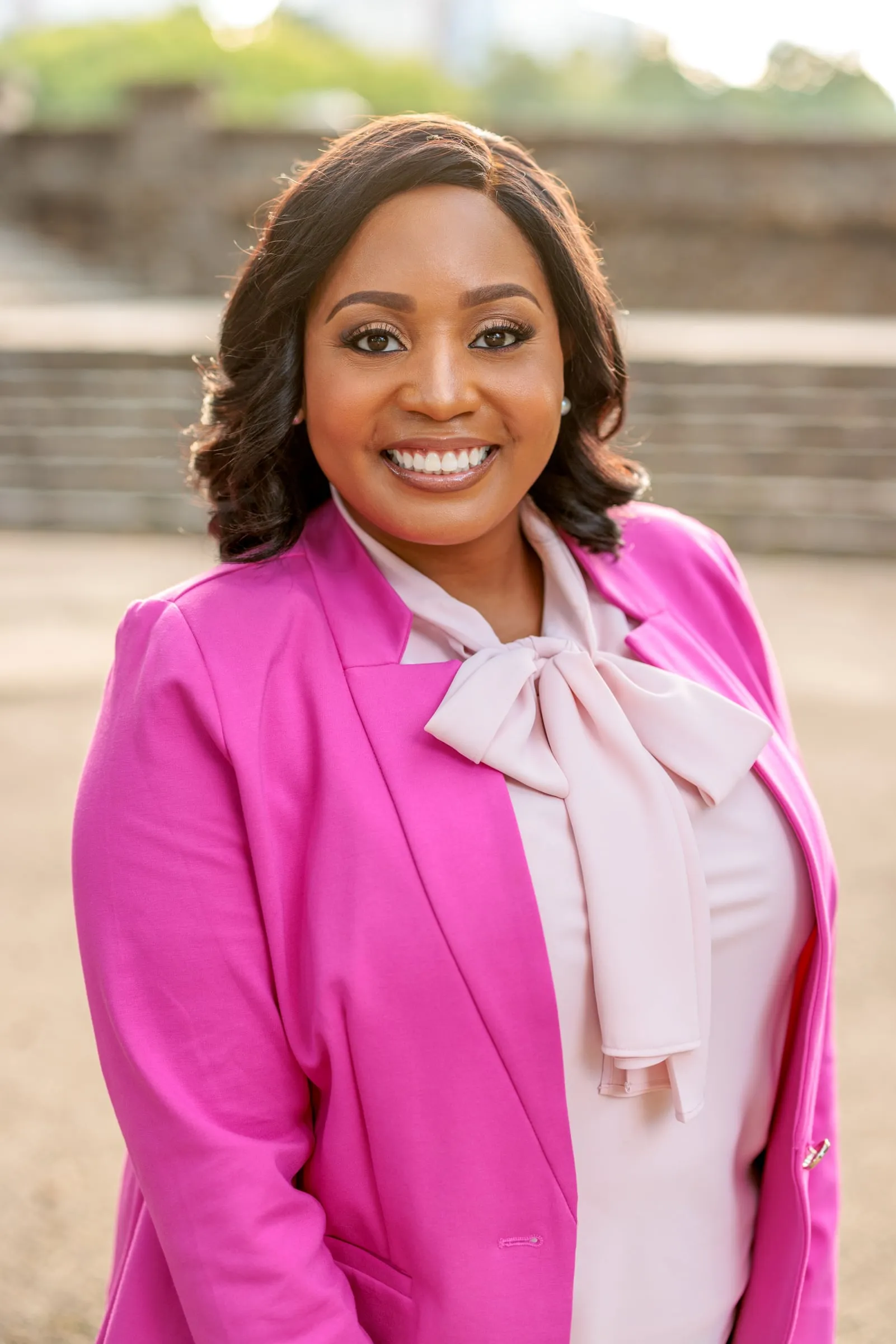 An outdoor headshot of a woman in a bright pink blazer and bow blouse on steps.