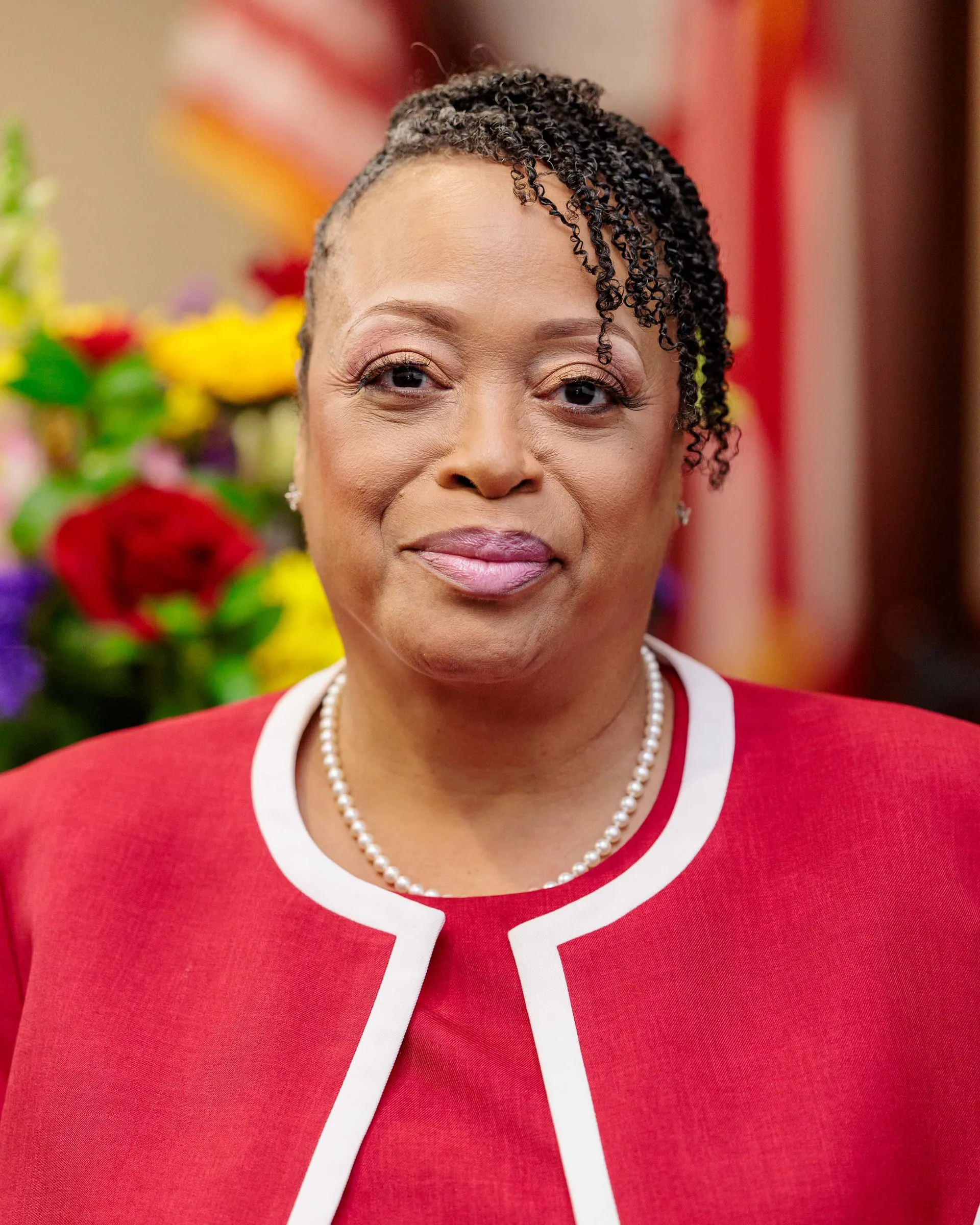 A woman in a red suit with white trim and pearls before flowers and a flag.