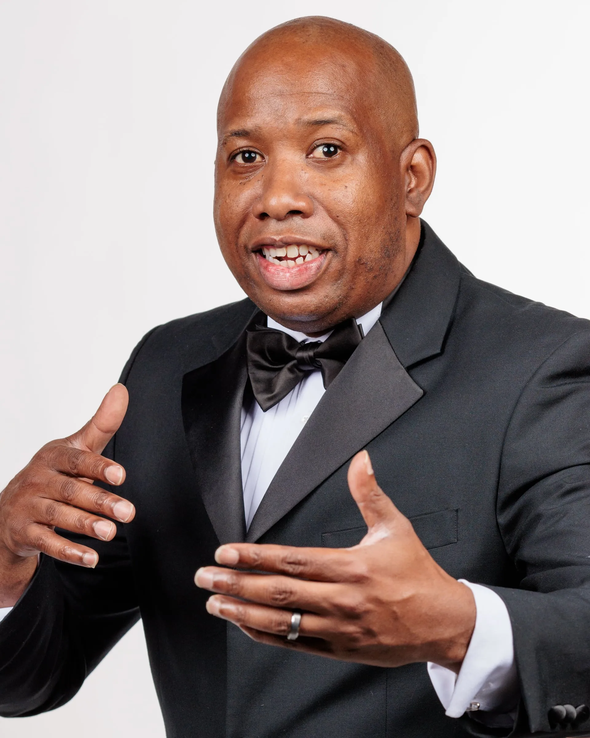 A man in a black tuxedo and bow tie mid-gesture as he speaks, on white.