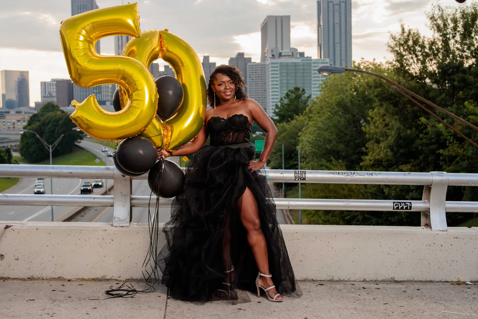 A woman in black tulle holding gold 50 balloons on a bridge above the Atlanta skyline.