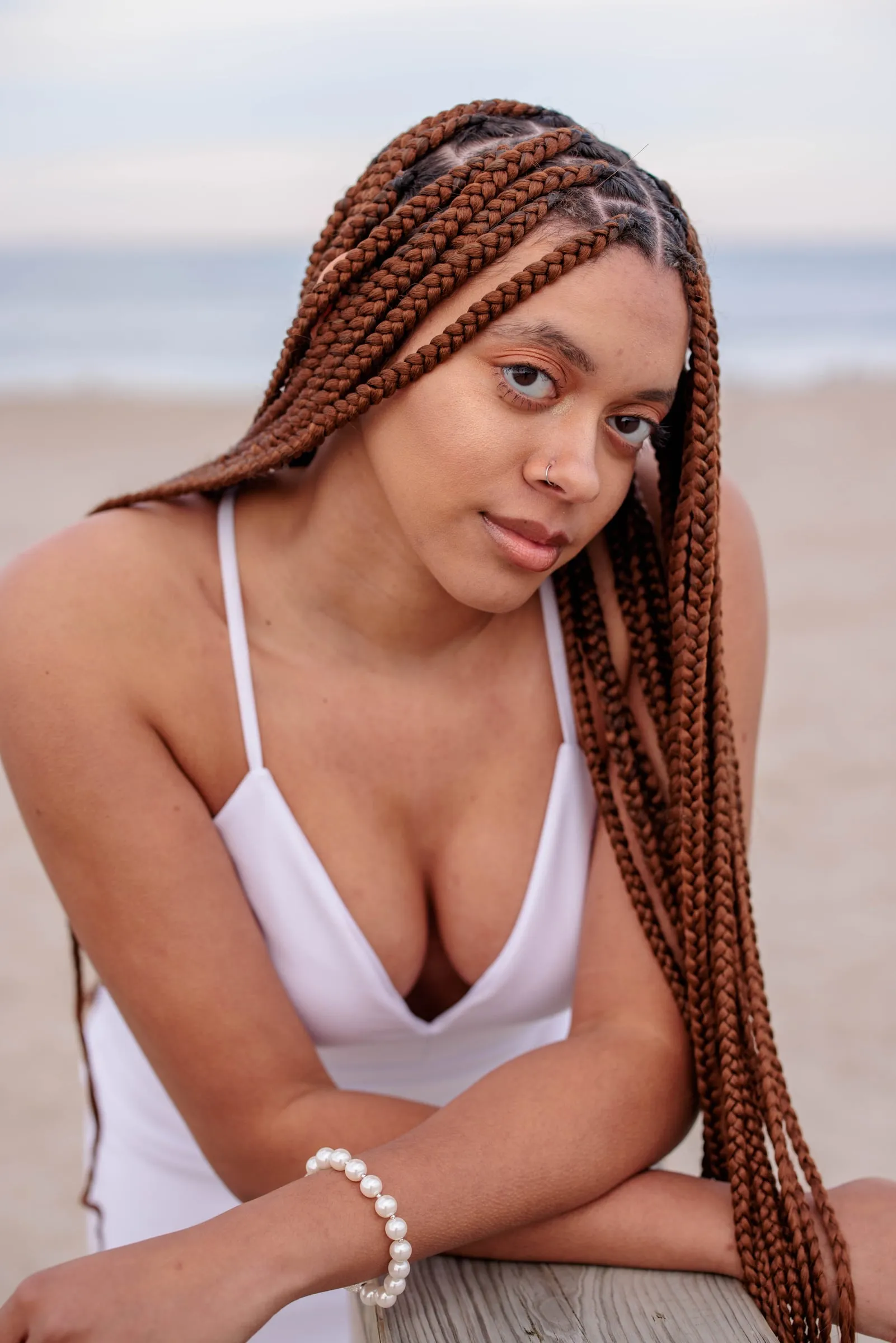 A young woman with long braids in a white top leaning forward on a beach boardwalk.
