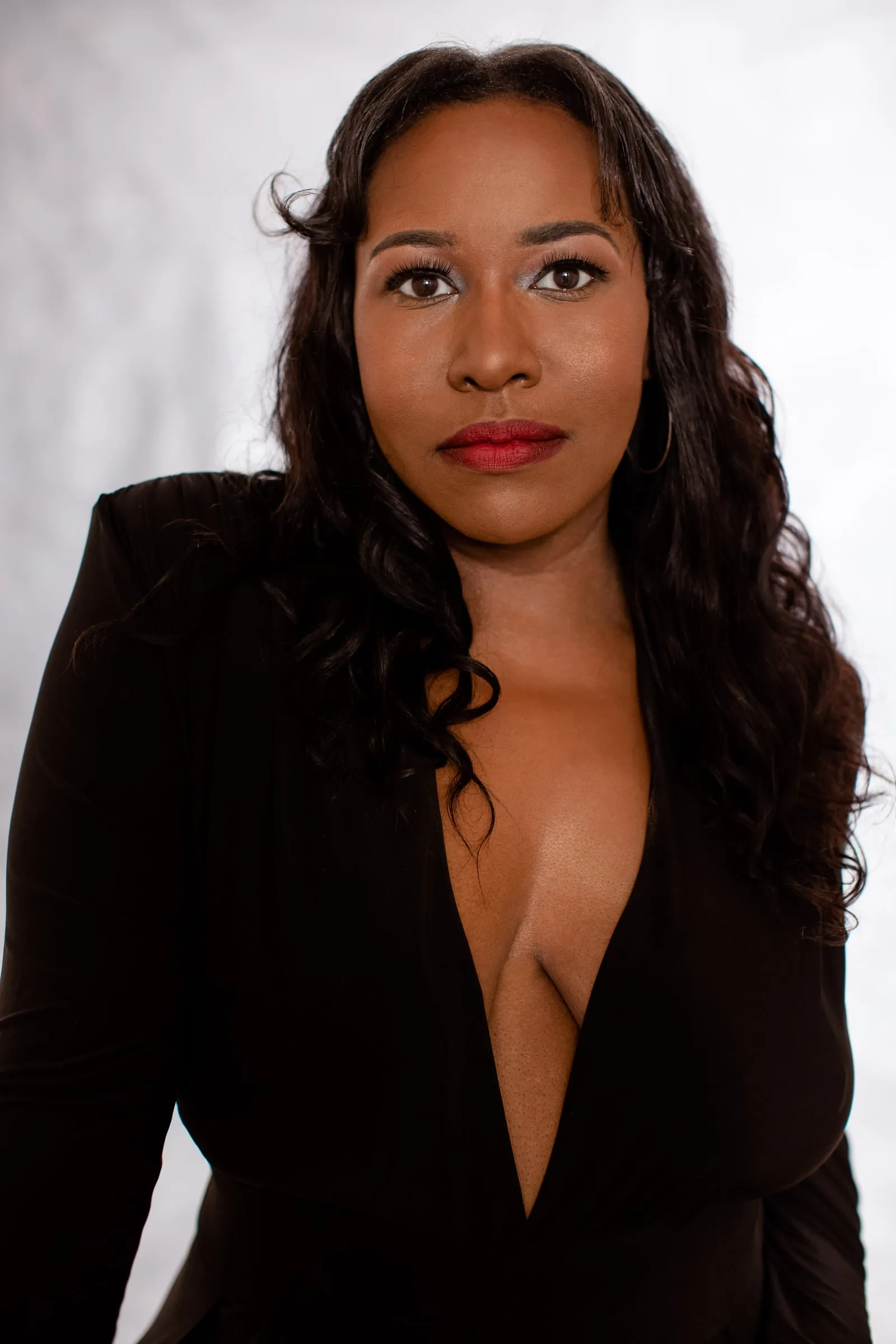 A woman in a plunging black dress with loose waves and red lips on grey.