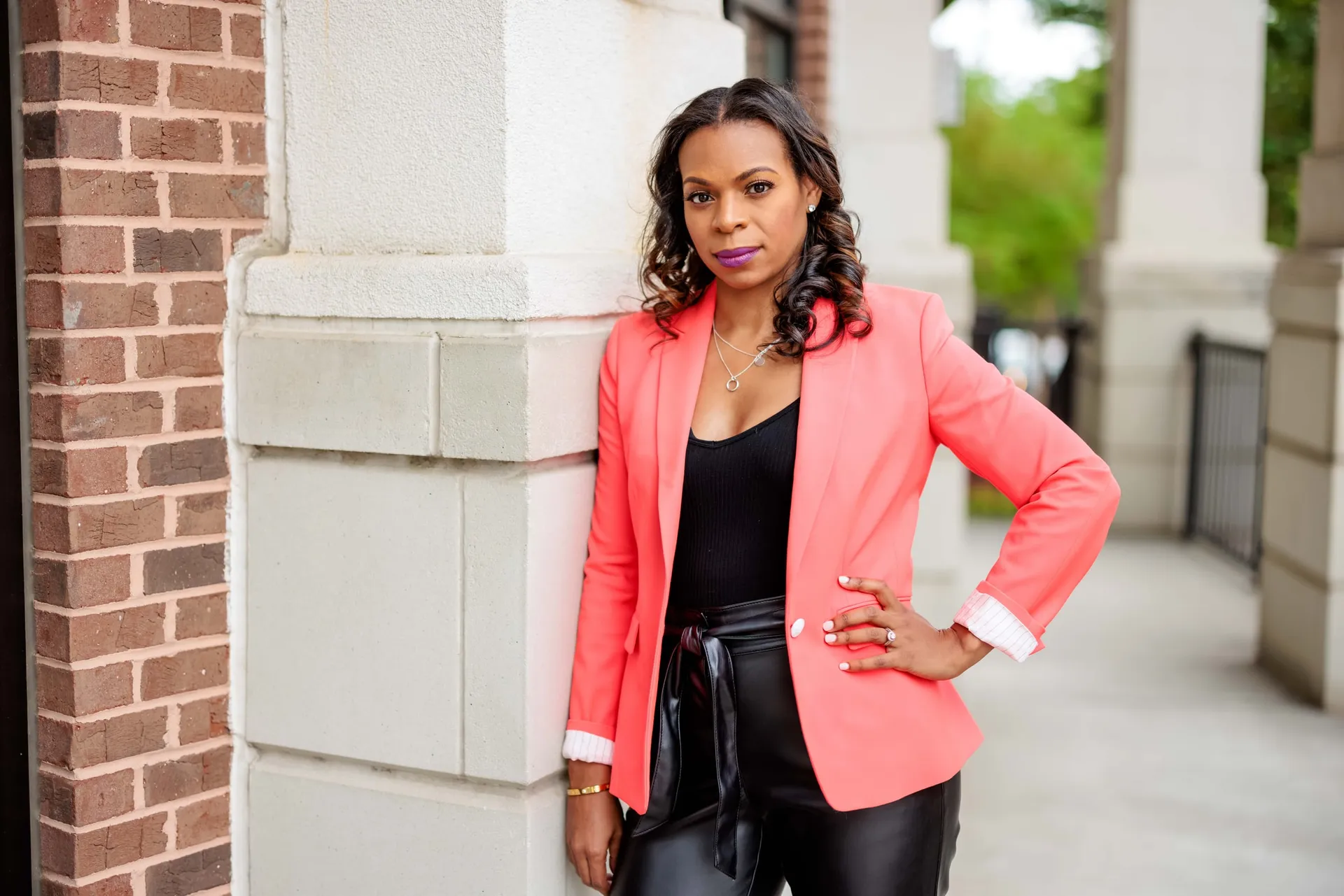 A woman in a coral blazer and leather trousers leaning on a stone column.