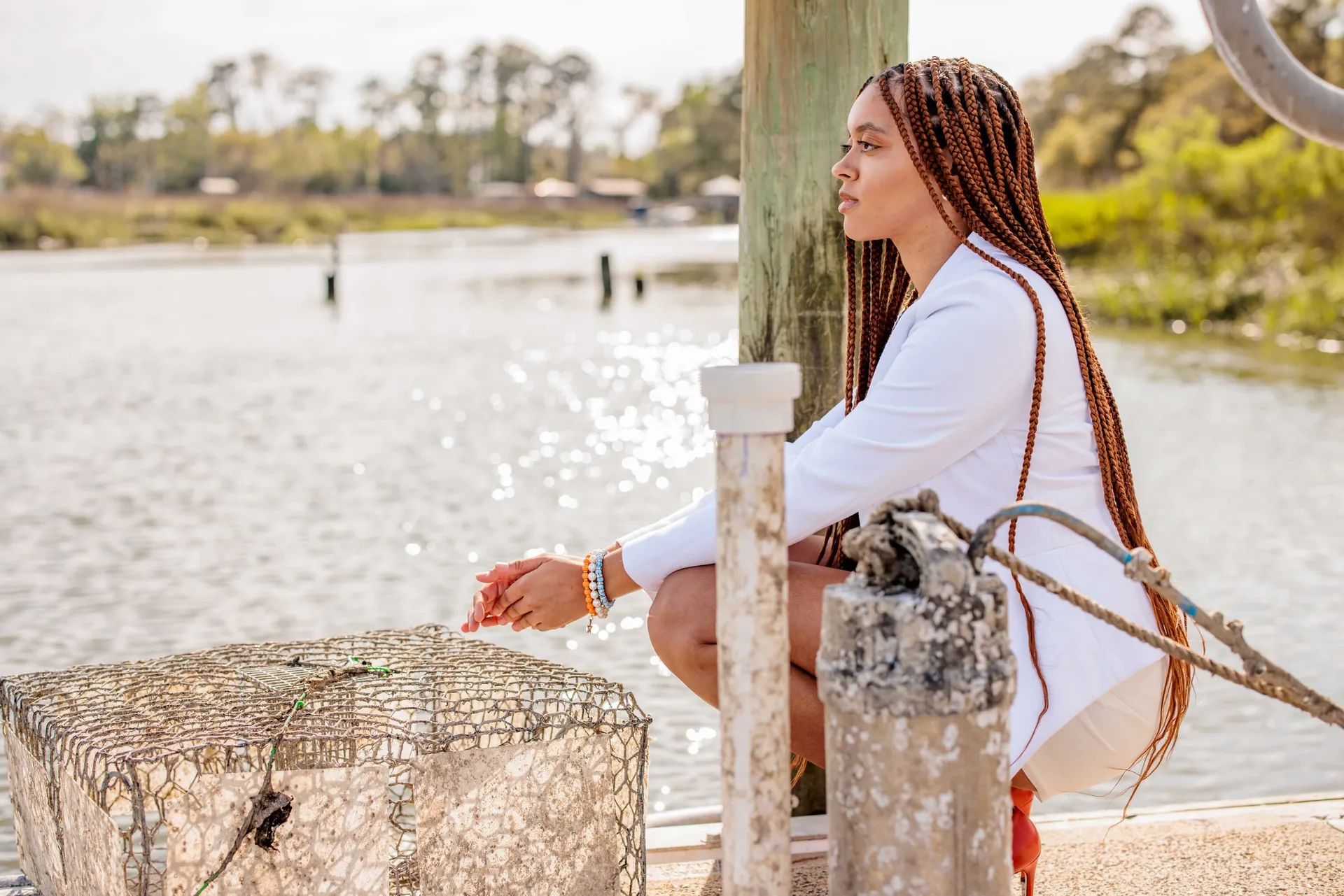 A young woman in white crouched on a dock beside a crab trap, looking seaward.