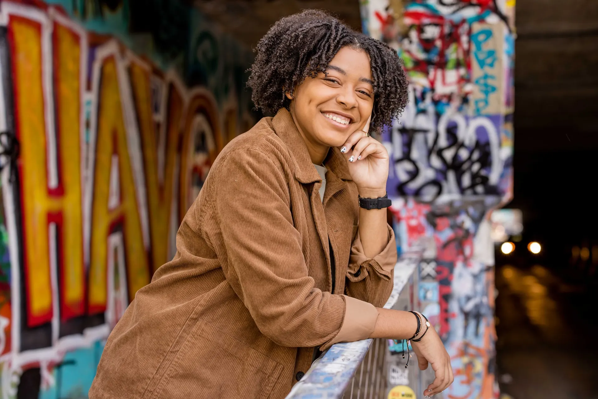 A senior in a tan corduroy jacket smiling against a graffiti-covered tunnel railing.