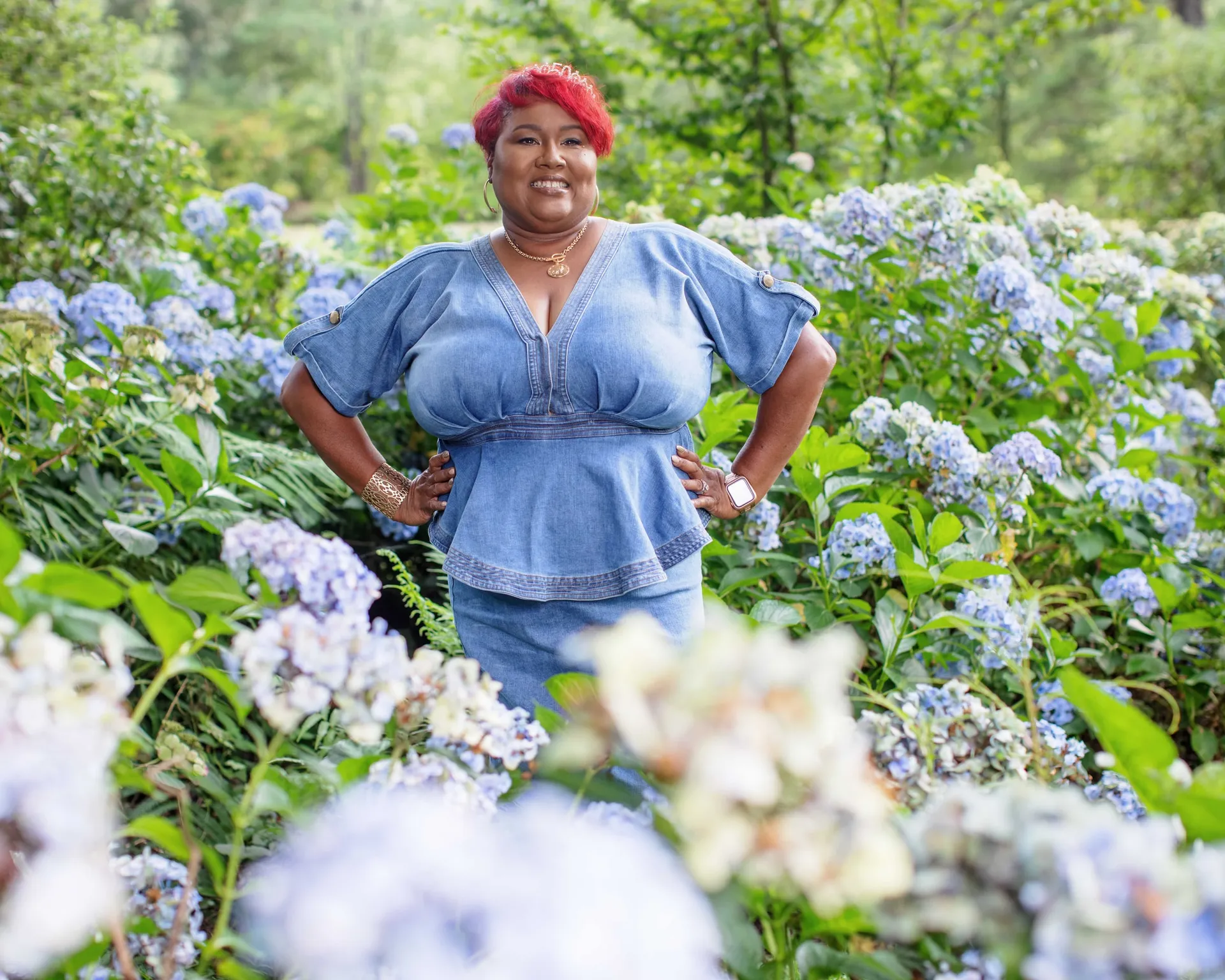 A woman with red hair in a denim top among blue hydrangeas, hands on her hips.