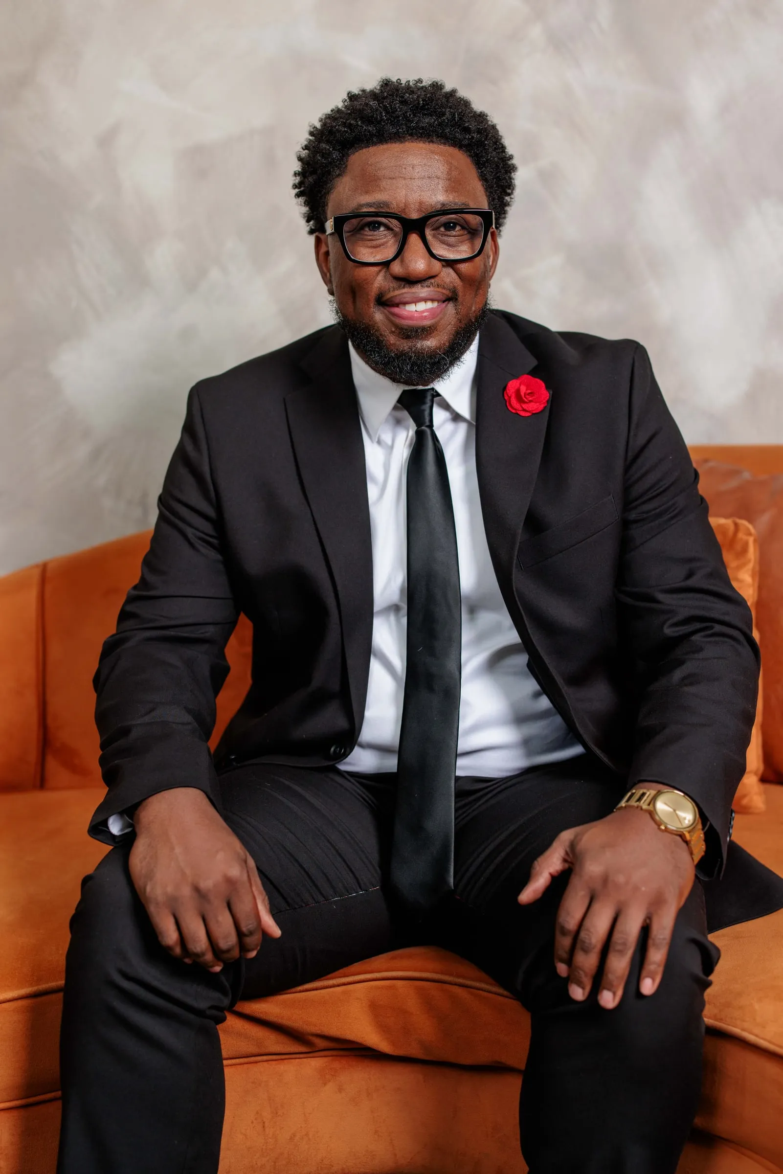 A man in glasses and a black suit with a red boutonniere laughing on orange velvet.