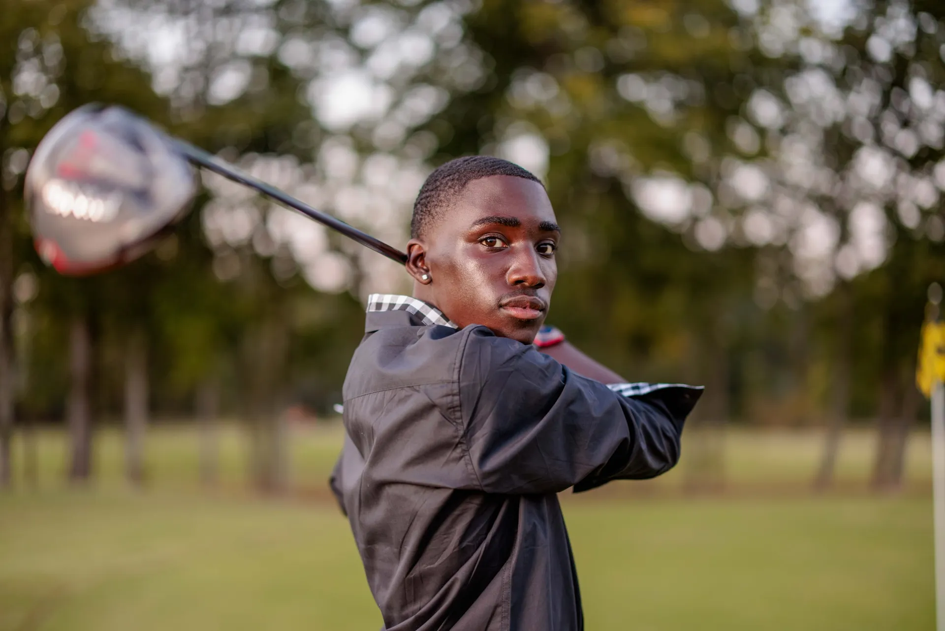 A senior in a black jacket following through a drive on a tree-lined golf course.