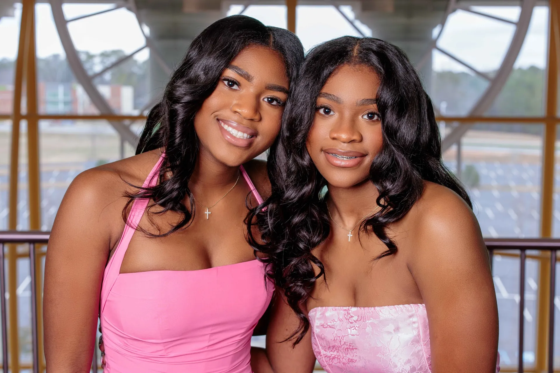 Two sisters in pink dresses smiling cheek to cheek on a glass-walled stairwell.